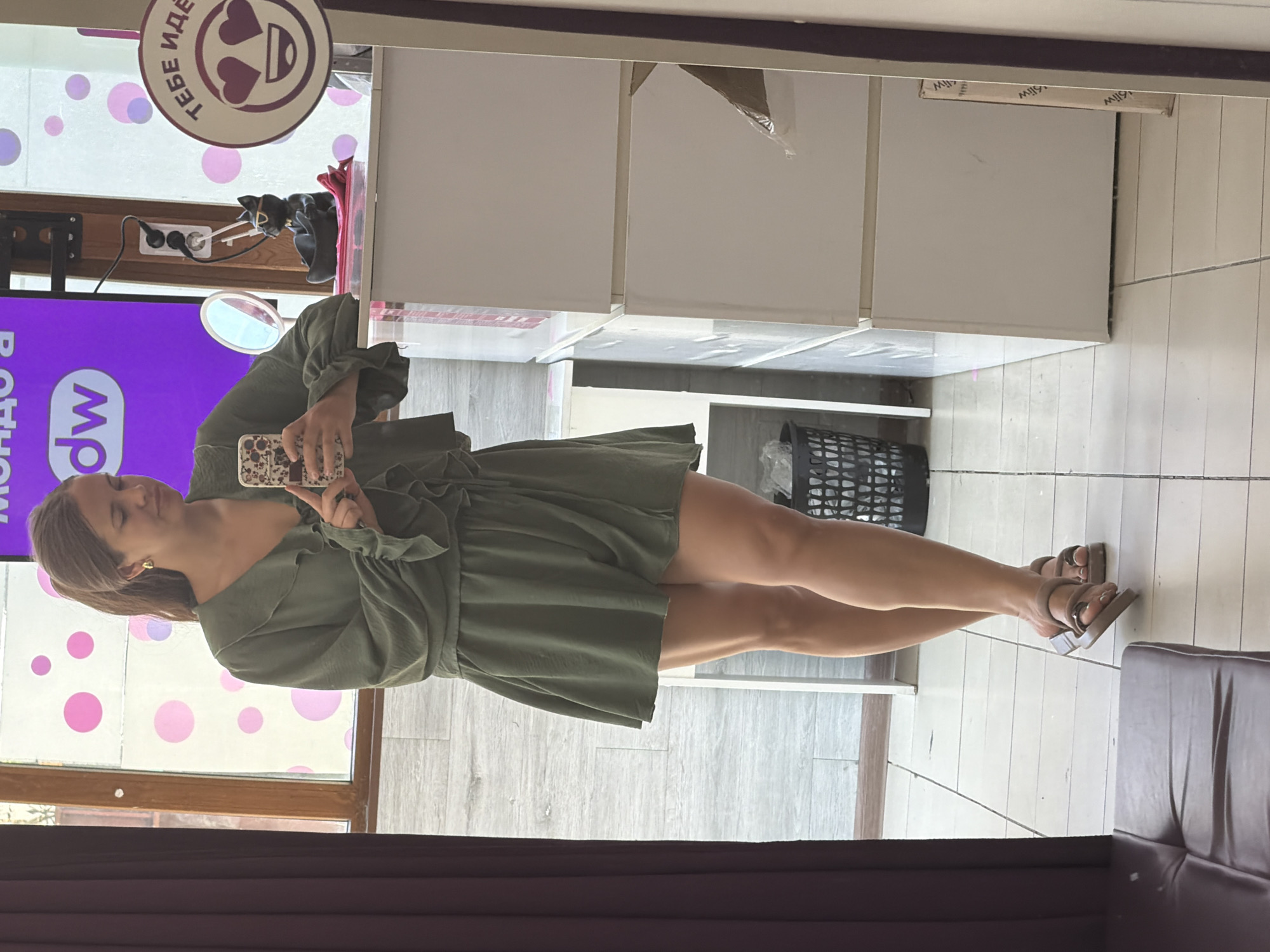 A young Caucasian woman with blonde hair tied in a ponytail is standing and taking a mirror selfie in a fitting room. She is wearing a green ruffled dress and brown sandals. She is holding a mobile phone in her right hand. In the background, there are several white cabinets, a trash can, and a window with pink polka dots on it. A sign with the Russian text "ТЕБЕ ИДЁТ" which translates to "It suits you" and a heart-shaped emoji is visible on the right side of the image. The woman appears to be in a clothing store or boutique. The environment is indoors with tiled flooring. The overall scene suggests a casual shopping experience.