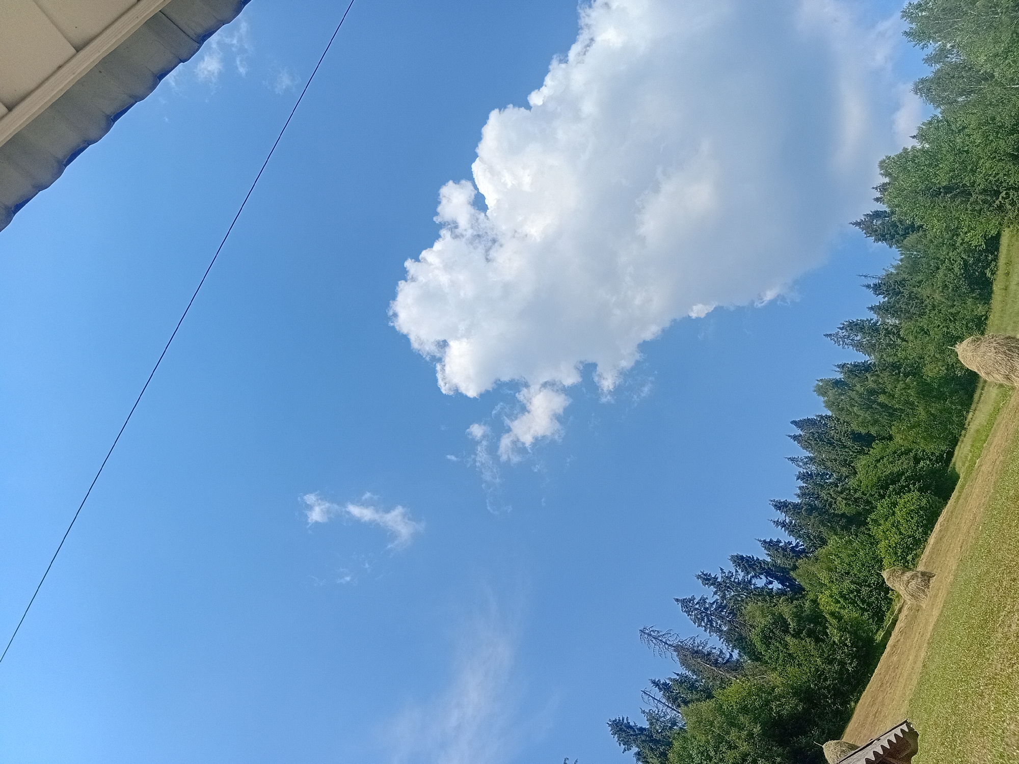The image depicts a rural scene with a bright blue sky dominating the upper portion. A few wispy white clouds are scattered across the sky, with a larger, more substantial cloud formation on the right side of the frame. A thin, dark line, possibly a wire or cable, stretches diagonally across the sky from the top left to the top right. The lower portion of the image shows a grassy hillside with several haystacks. A cluster of coniferous trees forms a dense treeline along the top of the hill. A portion of a building's roof structure is visible in the bottom left corner. The environment suggests a sunny day in a natural, possibly agricultural, setting. The location is specified as the city of Verkhniachka, Ukraine. No people or specific actions are visible in the image.