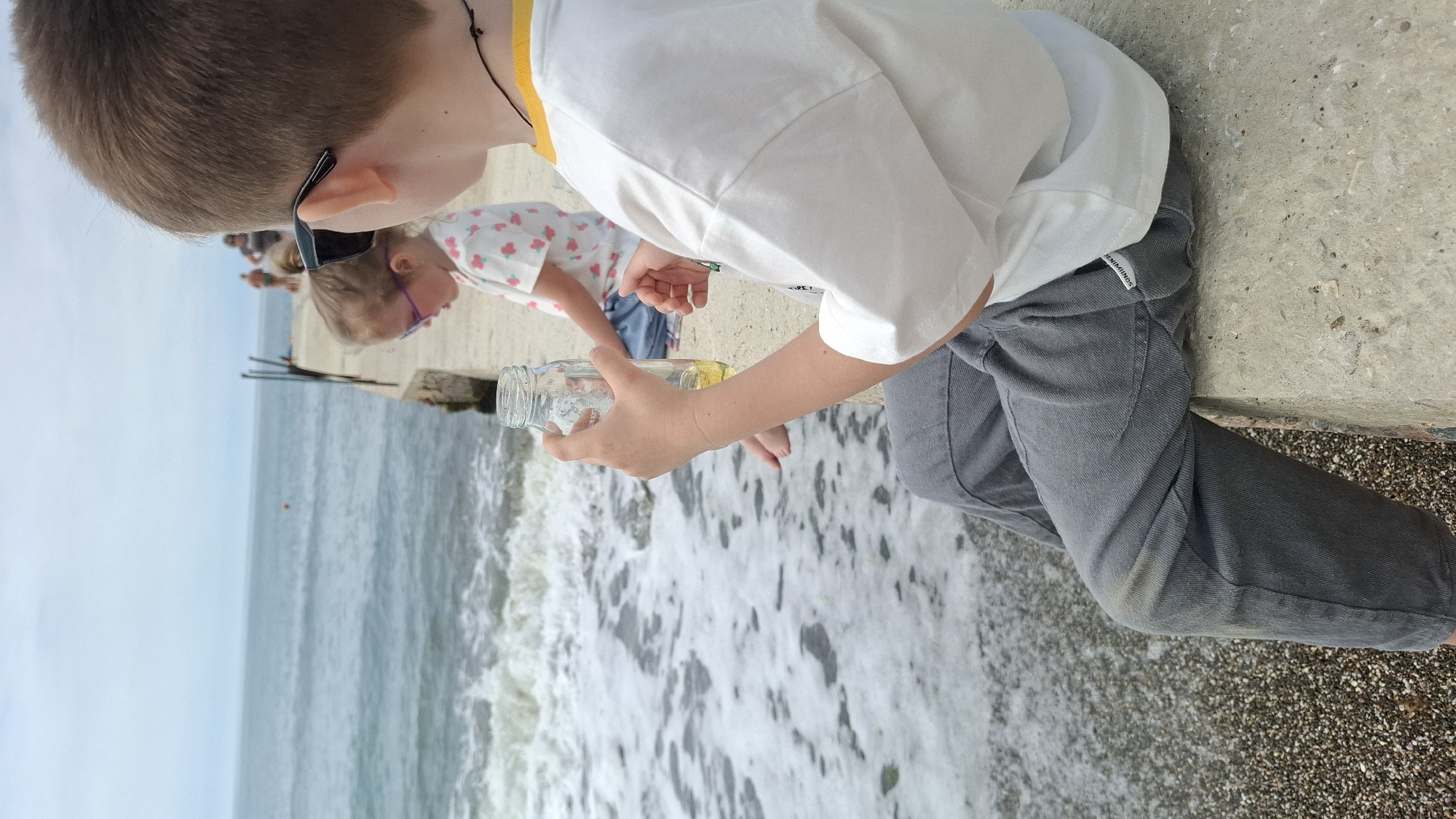 Two children are positioned on a concrete pier or seawall next to a body of water, likely the ocean, under an overcast sky. The image is taken from a slightly elevated perspective.
In the foreground, a boy with light brown hair is seen from the side, looking down towards the water. He wears black sunglasses pushed up on his head/ear, a white short-sleeved t-shirt with a yellow trim around the collar, and dark grey pants. His right hand holds a clear glass bottle that contains a yellowish liquid, possibly oil, floating on top of a clear liquid, presumably water.
Behind and slightly to the left of the boy is a younger child, likely a girl, also looking down. She has light hair pulled back in pigtails and wears purple-framed glasses and a white top with a scattered, colorful floral pattern.
The sea stretches into the distance, appearing choppy with white foam from waves breaking near the pier. A small red buoy is visible far out on the water, and in the distance, several blurred figures are on what looks like a continuation of the pier. Wooden poles protrude from the water further along the pier. The children seem focused on an activity involving the bottle and the water.
On the boy's grey pants, near the left hip pocket, a white label with the text "SIMMINDS" is partially visible. The overall impression is one of calm observation or interaction with the natural environment.