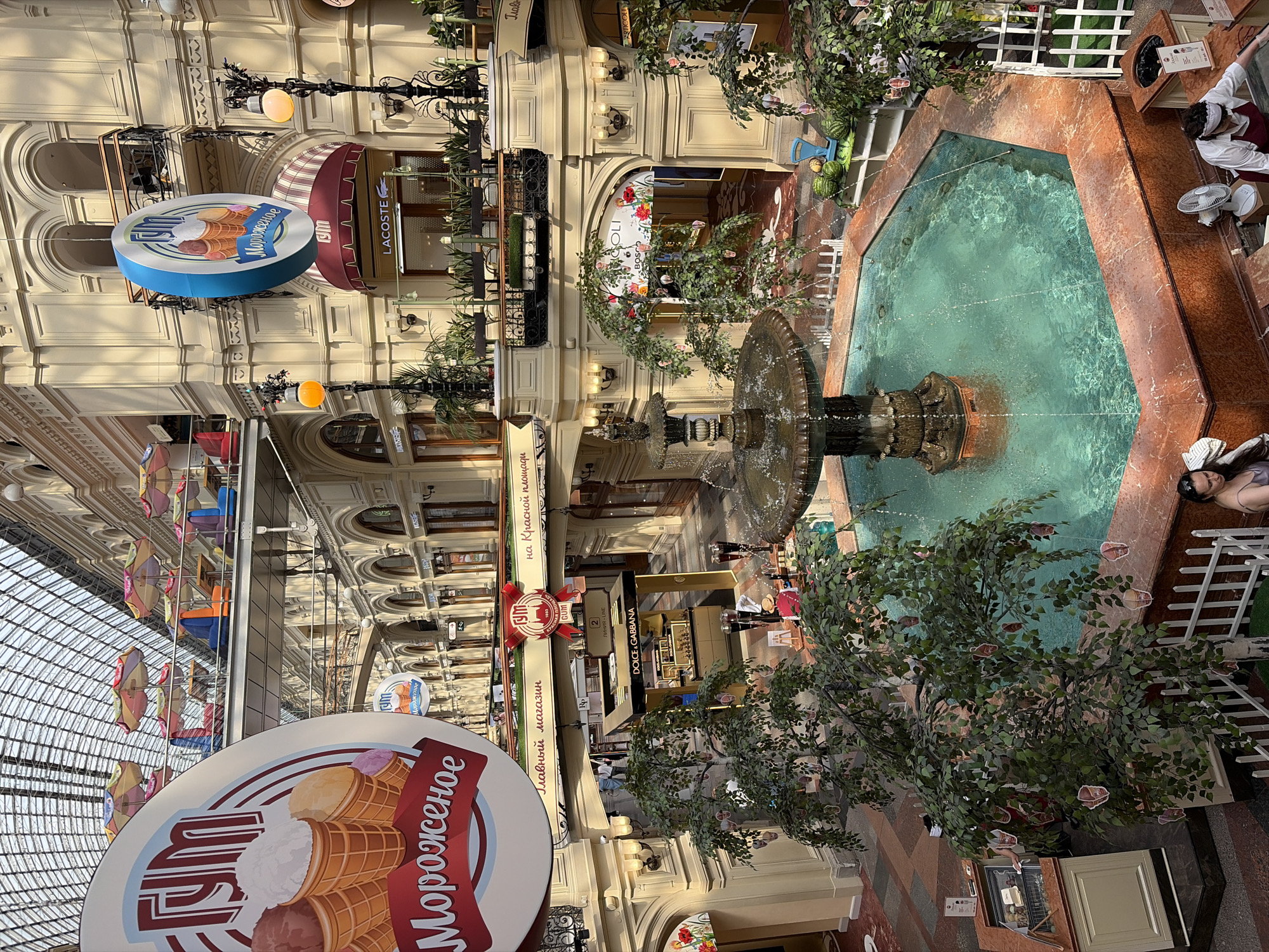 The image captures an interior view of GUM, a famous department store located on Red Square in Moscow, Russia. The grand architecture features a glass-paneled ceiling, cream-colored walls with arched windows and balconies, and multiple levels connected by walkways. In the foreground, a multi-tiered fountain with turquoise water is the central focus. Lush artificial trees with decorative elements hang from their branches are placed around the fountain.
Several shops are visible on different levels, including Lacoste and Dolce & Gabbana. Large circular signs advertising ice cream from "ГУМ" (GUM) are prominently displayed, one in the upper left and another hanging from the ceiling. People are seen browsing at the shops and near the fountain, including a woman in the lower right wearing a white shirt and apron, appearing to be working. The overall atmosphere is bright and airy, suggesting a lively shopping environment.