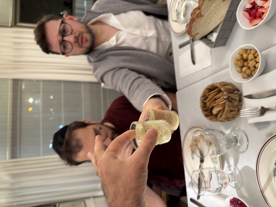 Two men at a table with a white tablecloth toast with glasses of light-colored liquid. One man wears glasses and a gray cardigan over a white shirt. The other man wears a maroon shirt and a black yarmulke. The table is set with bread, a small bowl of strawberries, a bowl of chickpeas, and a woven basket with round crackers. The background includes a window and partially visible decorations.
