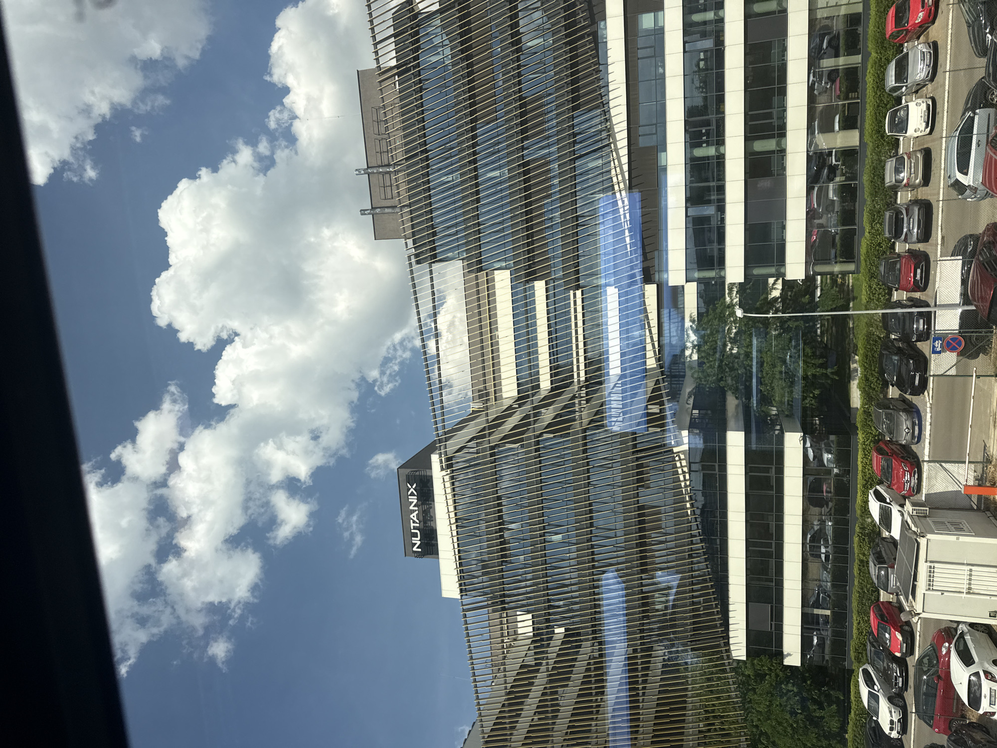 The image shows the Nutanix office building in Belgrade, Serbia. The building is a modern glass structure with horizontal louvers. The sky is blue with scattered white clouds. A parking lot with several cars is visible to the side of the building. A lamppost is also present. The image appears to have been taken from inside a vehicle due to the presence of a dark frame on the left side.