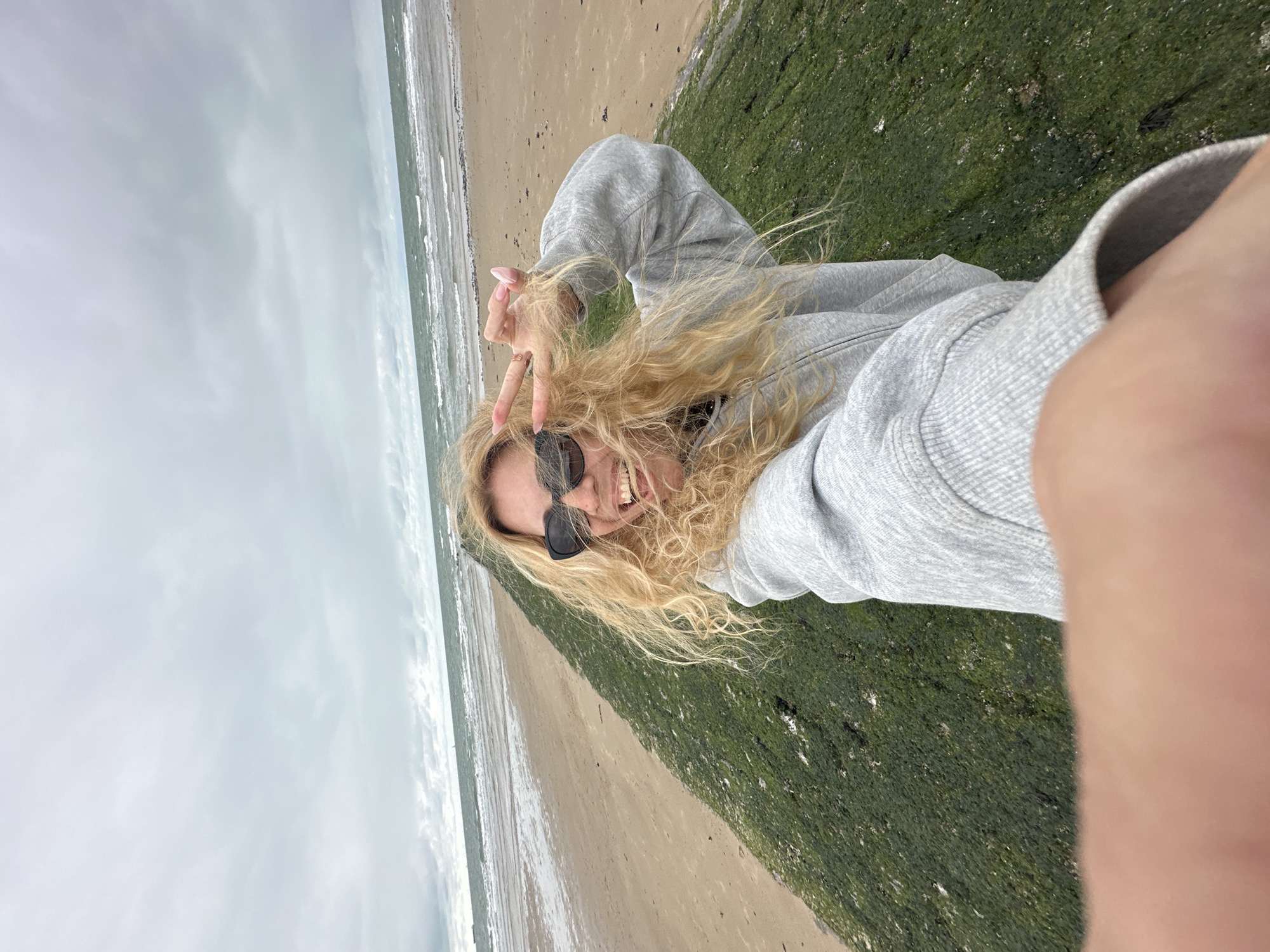 A young woman with long, wavy blonde hair and sunglasses smiles at the camera, making a peace sign with her fingers. She is wearing a grey hoodie. She appears to be on a beach, with the ocean and a sandy shore visible behind her. To her right, a section of green seaweed or algae covers a rocky or concrete structure. The sky is overcast, suggesting a cloudy or possibly stormy day. The lighting is diffused, indicating it might be daytime but not particularly sunny. The overall mood of the image is cheerful and relaxed, with the woman's expression conveying happiness and playfulness. There is no visible text in the image. The location context of Knokke-Heist, Belgium, suggests a coastal setting in the North Sea.