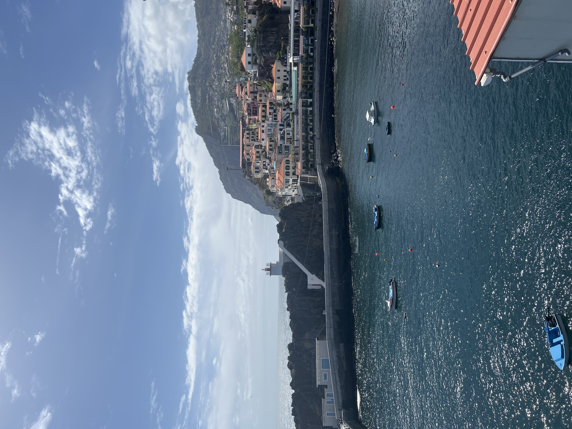 The image shows a view of Câmara de Lobos, Portugal, from a high vantage point. The landscape includes mountainside buildings, a coastal road, the sea, and the sky with scattered clouds. Several boats are in the water. In the foreground, the corner of a red-roofed structure is visible.