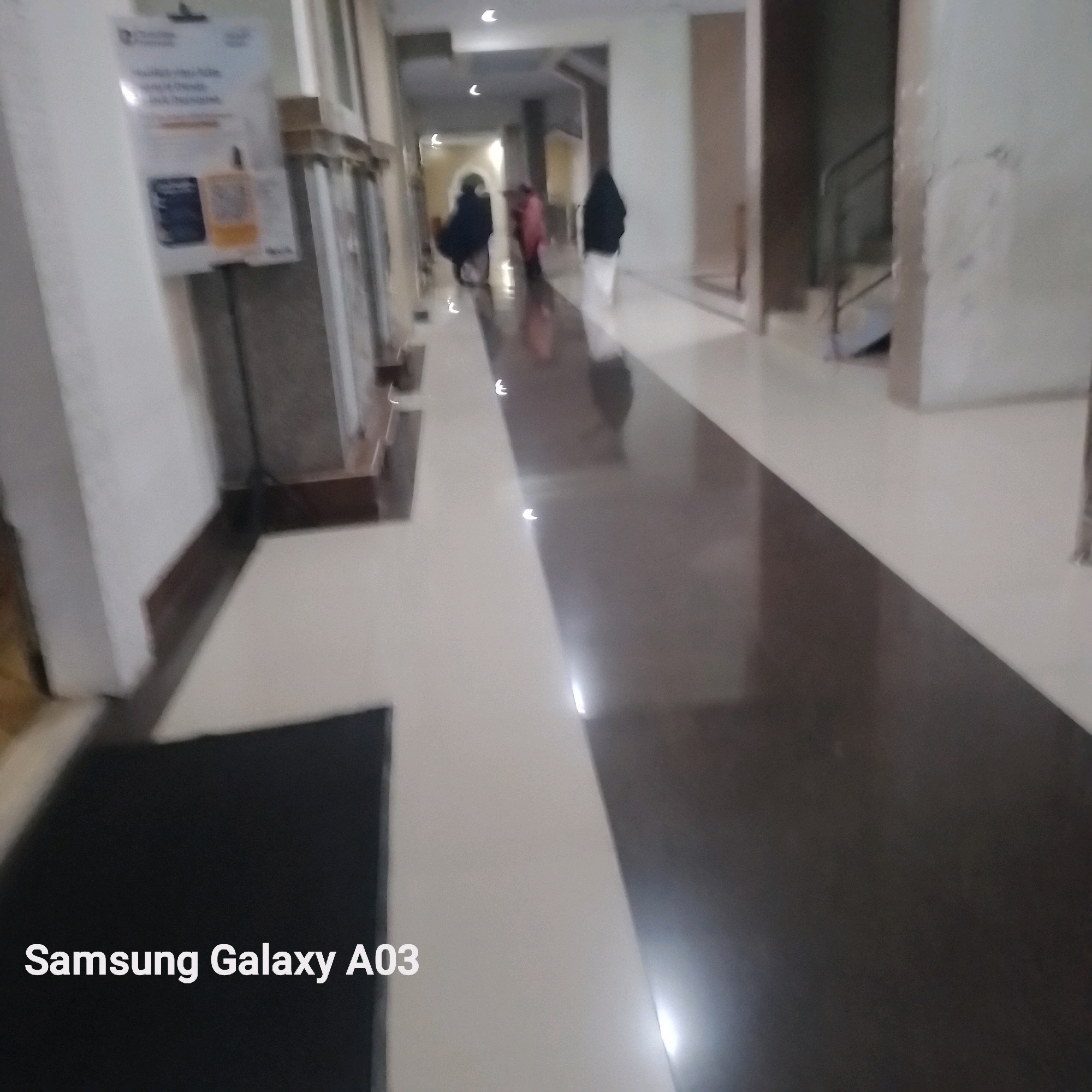The image depicts a blurred interior hallway, likely within a public building in Kenanga, Indonesia. The hallway features a polished, reflective floor with a distinct color contrast, with a lighter section on the left and a darker, glossy section on the right. Several indistinct figures, appearing to be people in modest attire, are visible walking down the hallway. One figure on the far left is wearing dark clothing, while a figure in the center is in a pink garment, and another on the right is in dark clothing with a white lower garment. The walls are a light color, and there are lights visible on the ceiling, casting reflections on the floor. On the left side of the hallway, a sign or poster is affixed to the wall, displaying text and what appears to be images of products. The lower left corner of the image contains a watermark that reads "Samsung Galaxy A03". The overall impression is of a mundane, everyday scene within an indoor space.