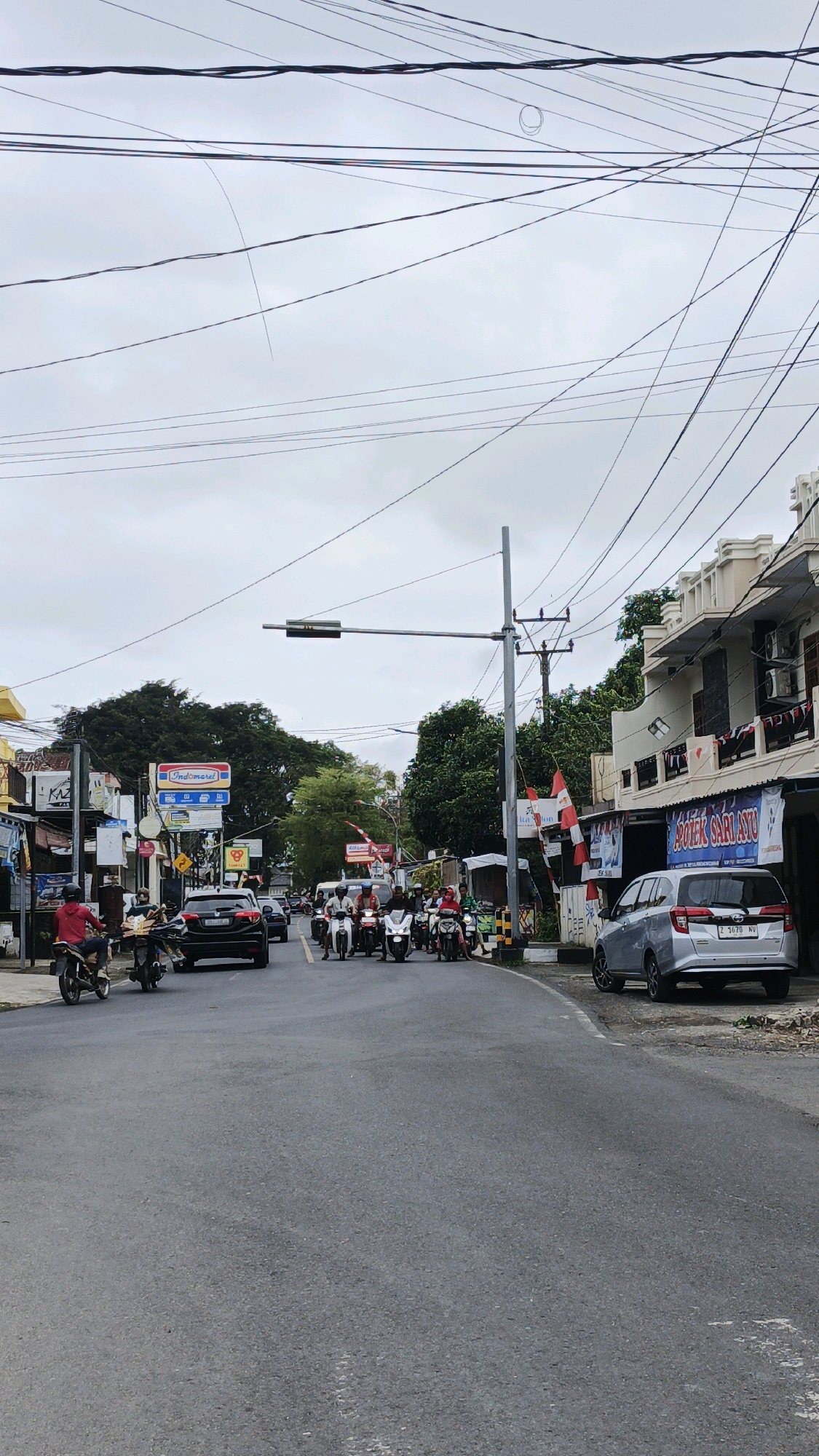 The image depicts a street scene in Desa Karangbenda, Indonesia, under an overcast sky. The road is paved and shows some signs of wear. On the right side of the road, there is a two-story building with a facade featuring white paint and decorative elements. A silver minivan is parked on the side of the road in front of this building, displaying the license plate "Z 1670 NV". A sign above a storefront on this building reads "APOTEK SARIAYU".
The left side of the road features several buildings, including a store with a prominent "Indomaret" sign. Various flags and banners are visible, some of which are red and white, possibly indicating national flags or decorations.
A traffic light is suspended over the intersection, with a traffic pole to its right. The road is populated with a number of motorcycles and a few cars. Several people are riding the motorcycles, some are waiting at the traffic light. One person on a motorcycle in the foreground on the left is wearing a red jacket. Another motorcycle in the middle ground on the left is black and is being ridden by a person in a white shirt. The overall atmosphere suggests a moderately busy intersection in a town or city. The background is dominated by lush green trees. Numerous overhead wires crisscross the sky.