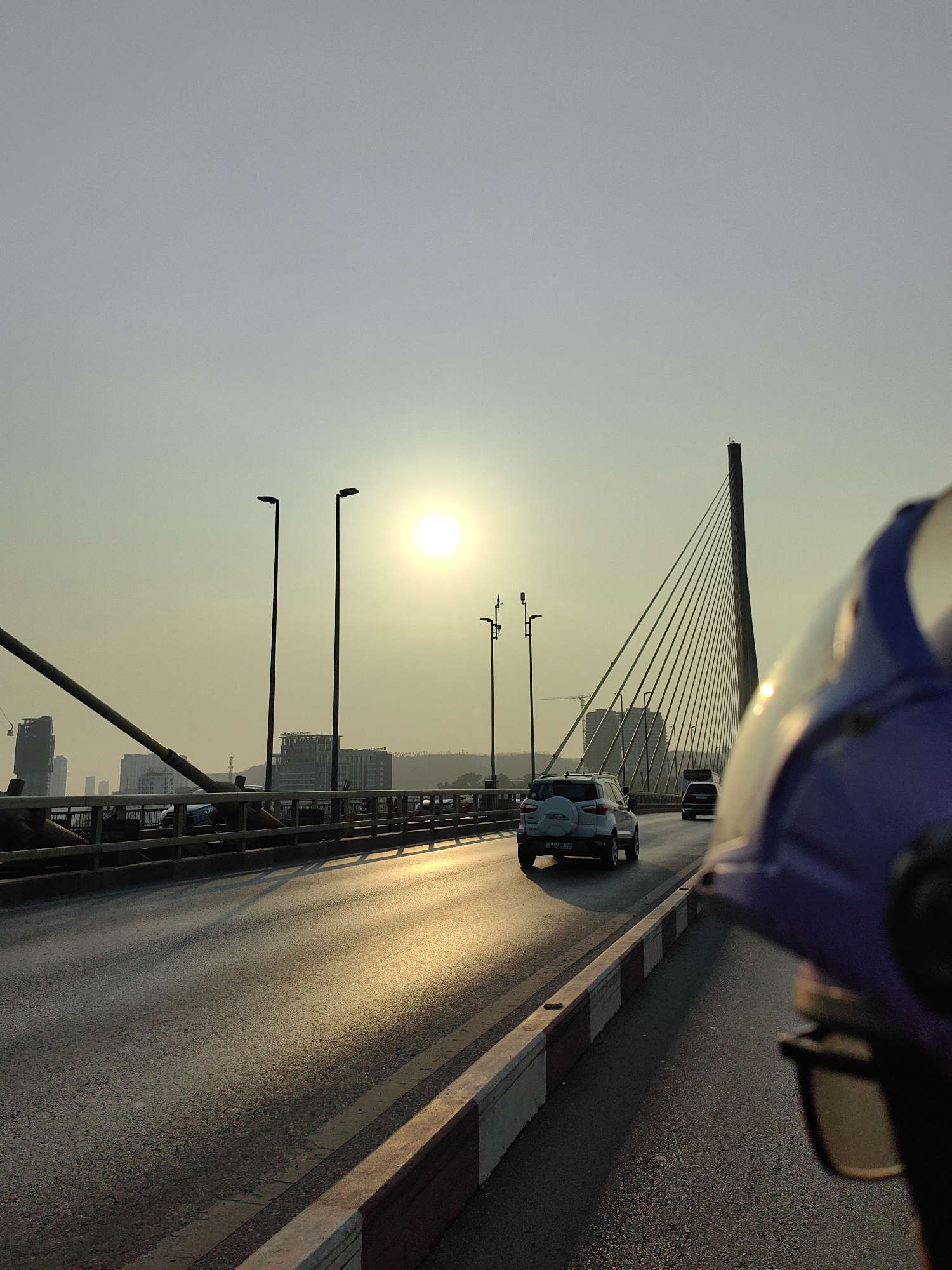 The image shows a view from a vehicle, likely a motorcycle, on a bridge in Hong Gai Ward, Vietnam. The sun is low in the sky, creating a hazy effect and casting long shadows. Several cars are on the bridge. A tall cable-stayed bridge pylon rises in the background. There are buildings in the distance. In the foreground, a blue helmet is visible. The road surface has a reflective sheen from the sun. The bridge has railings and street lights.