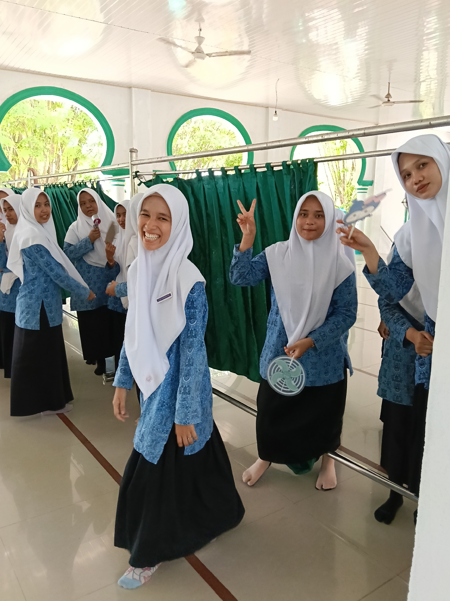 In Lhokseumawe, Indonesia, a group of women wearing white hijabs, blue and white patterned long-sleeved shirts, and black skirts are standing in a row inside a building with white walls and green-trimmed arched windows. Green curtains hang behind a metal railing. One woman in the foreground is smiling broadly, while another is holding up a peace sign. A third woman holds a small handheld fan. The floor is tiled and patterned. Ceiling fans are visible on the white ceiling.