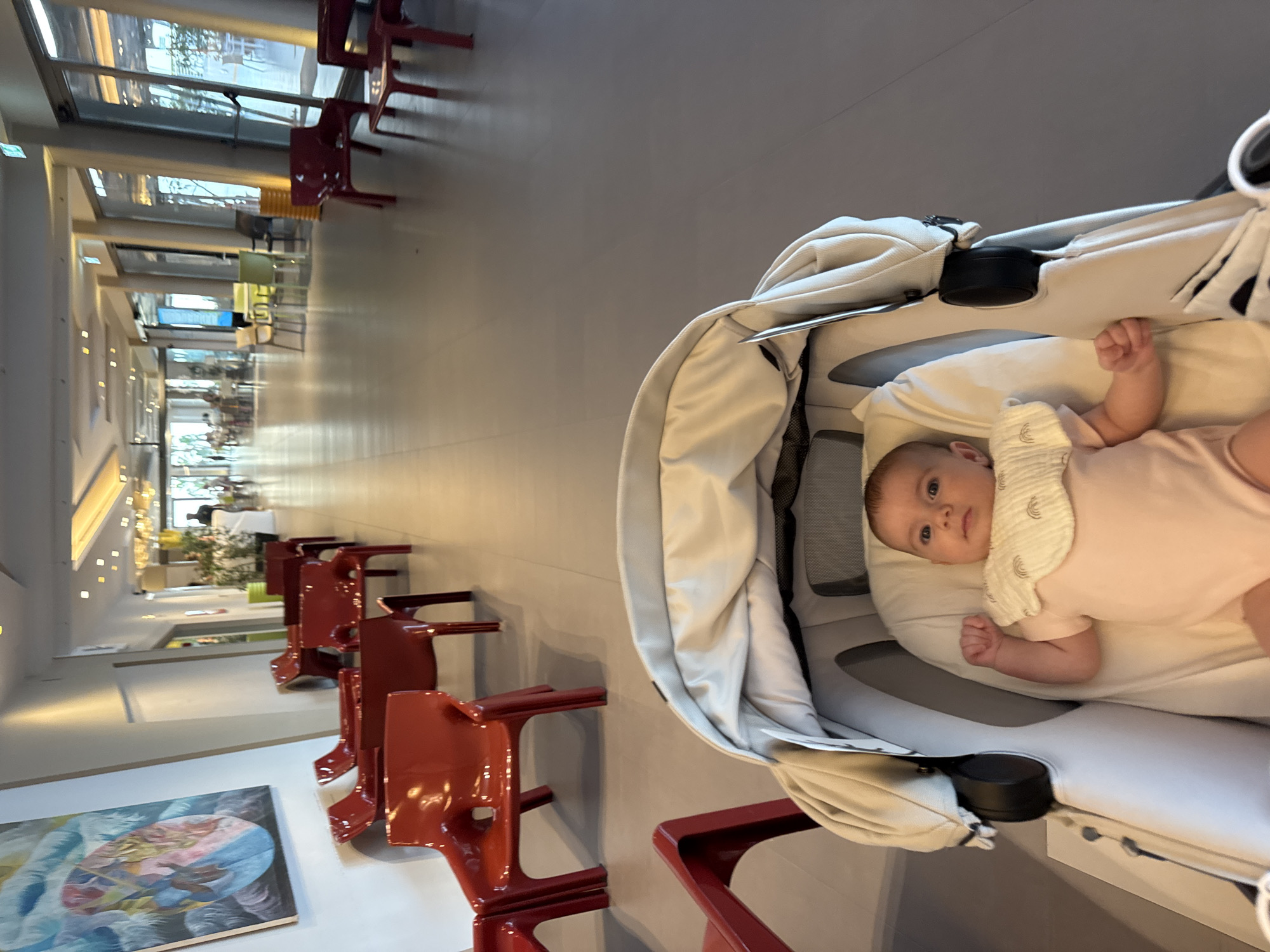 An infant lies in a cream-colored stroller, facing forward, with a bib around its neck. The stroller is positioned in a brightly lit, modern hallway. To the left, a colorful abstract painting hangs on a white wall, accompanied by a row of bright red chairs. The hallway extends into the distance, lined with windows and more seating areas, suggesting a public space like a hotel lobby, waiting area, or airport terminal. The floor is a smooth, gray surface. The overall impression is one of a clean, contemporary environment.