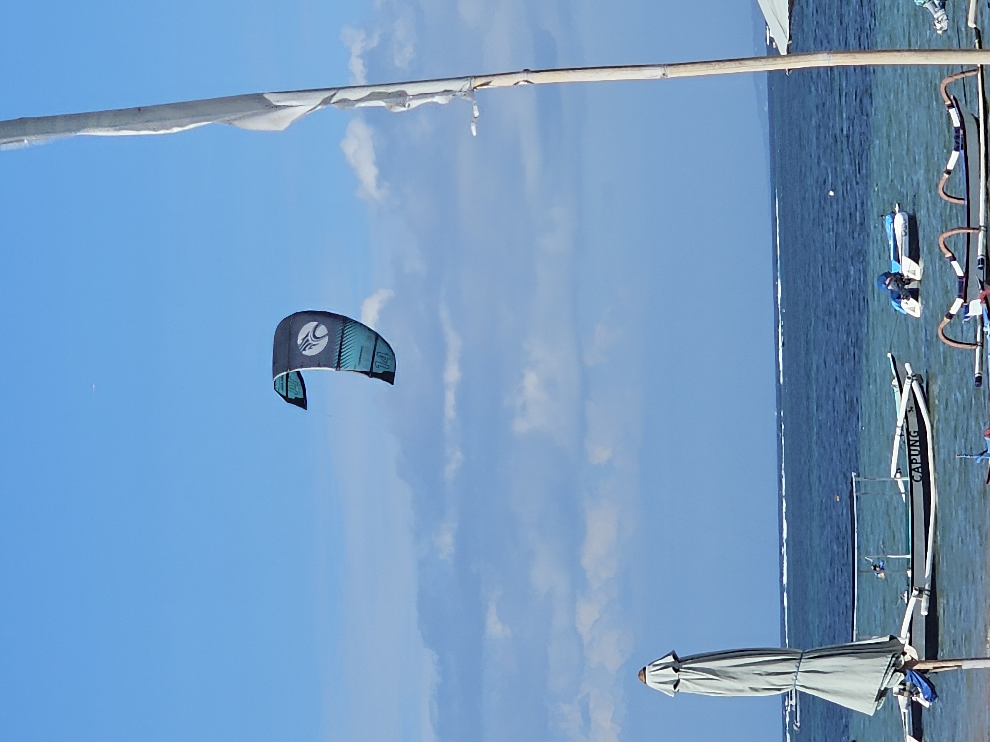 The image shows a beach scene likely in Denpasar, Indonesia. The view is tilted 90 degrees counter-clockwise. A black and turquoise kiteboarding kite is in the air against a blue sky with scattered clouds. The ocean is visible to the right, with several outrigger canoes on the beach. One canoe has "CAPUNG" written on its side. A beach umbrella is partially visible at the bottom of the image. A person with blue clothing and a white boat can be seen in the sea, by the boat.
