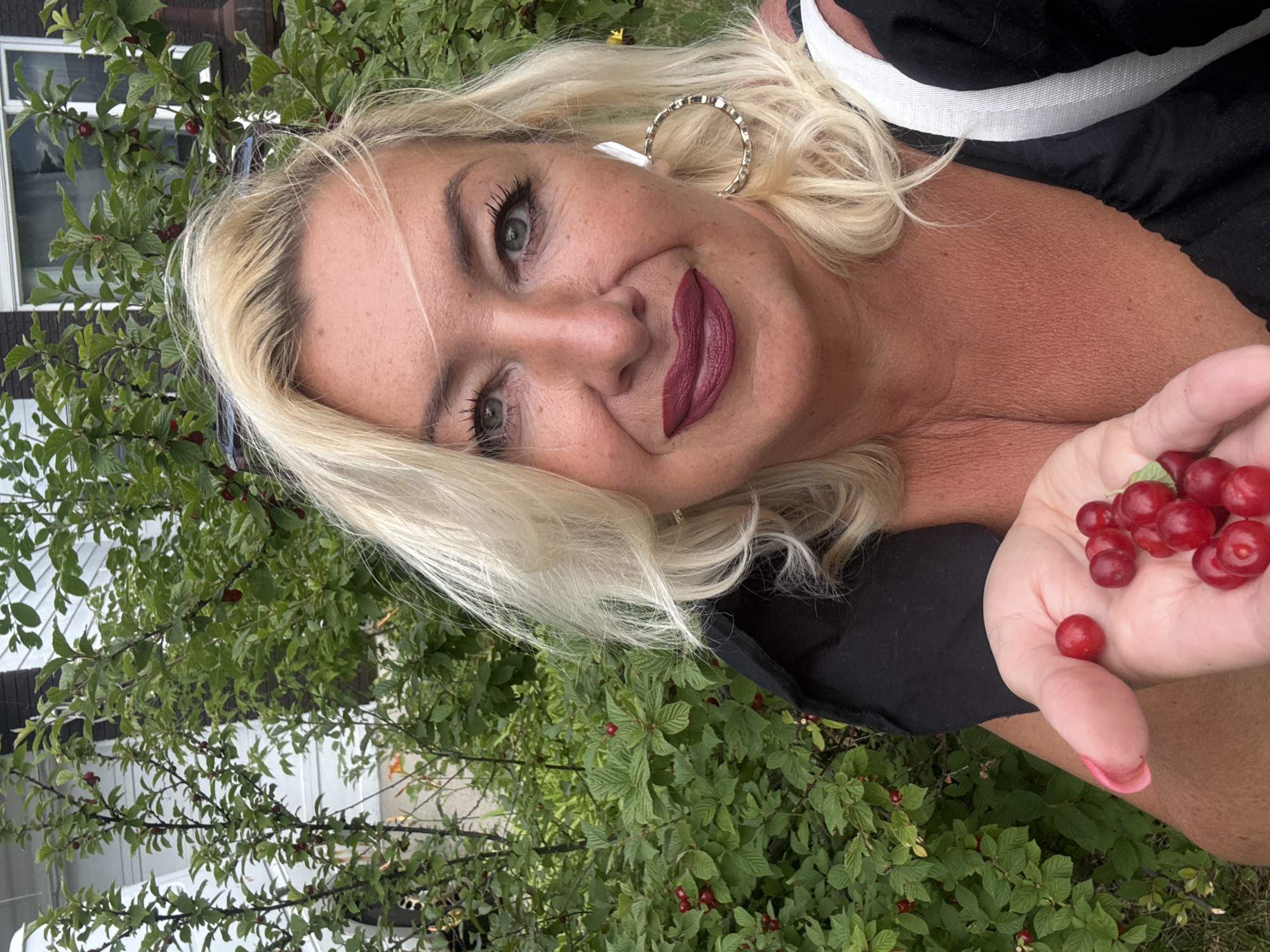 A woman with blonde hair and dark lipstick is holding a handful of red berries in her palm, with a bush full of berries in the background. She is wearing a black top. The scene appears to be outdoors, possibly in a garden or yard, with a house visible in the background. The image was taken in Calgary, Canada.
