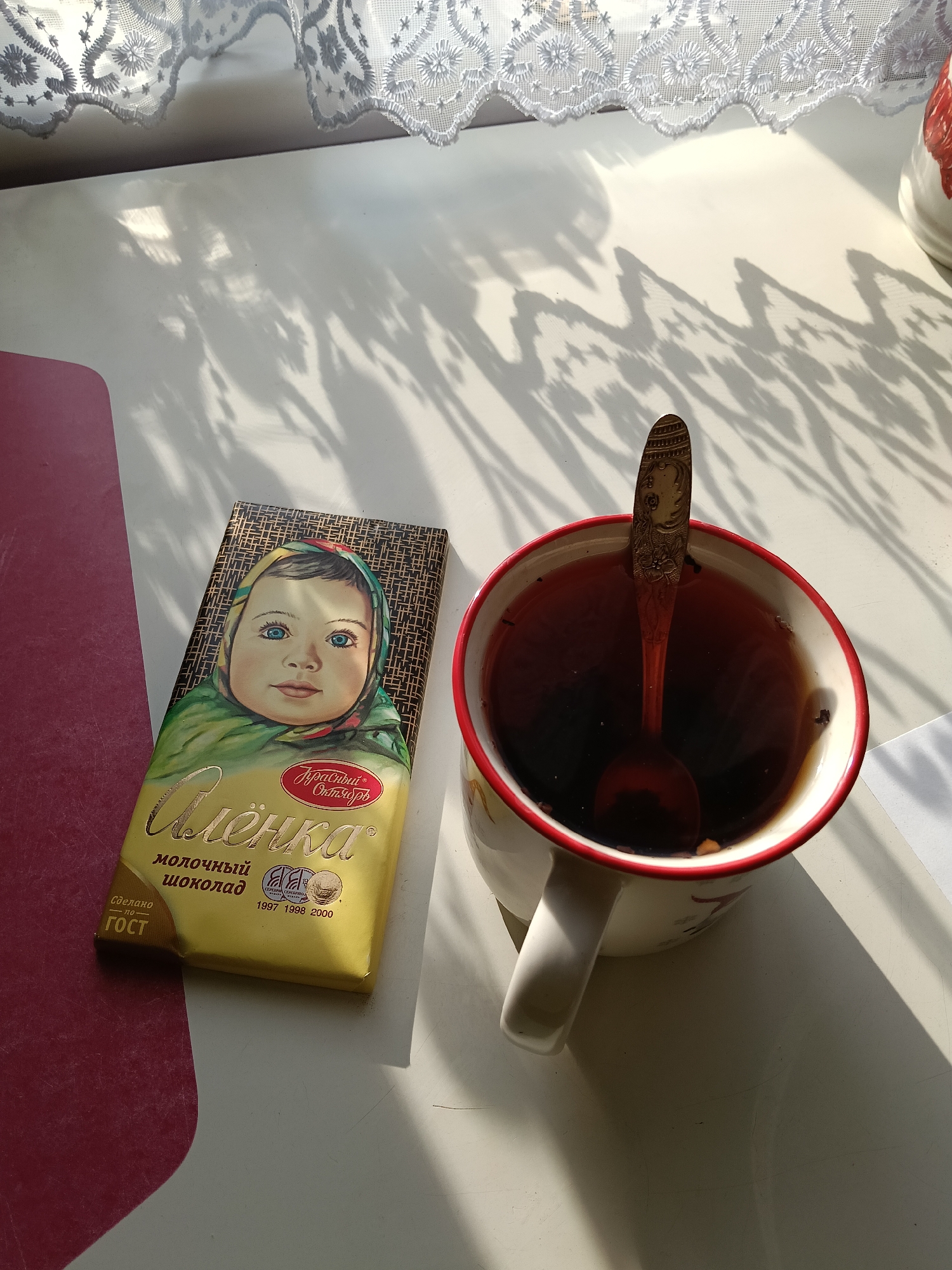 A still life scene captures a mug filled with a dark liquid, likely tea, and a spoon resting inside it. Beside the mug is a chocolate bar with the face of a child on its packaging, identified as "Alionka" milk chocolate, a popular brand in Russia. The scene is bathed in bright sunlight, casting shadows on the white surface of what appears to be a table. Part of a red cutting board is visible on the left side of the frame, and a lacy curtain is positioned at the top of the image. The scene suggests a domestic, possibly breakfast or tea time setting.
There is no visual information in this image that allows for the determination of location in the city of Красный Маяк, Russia.