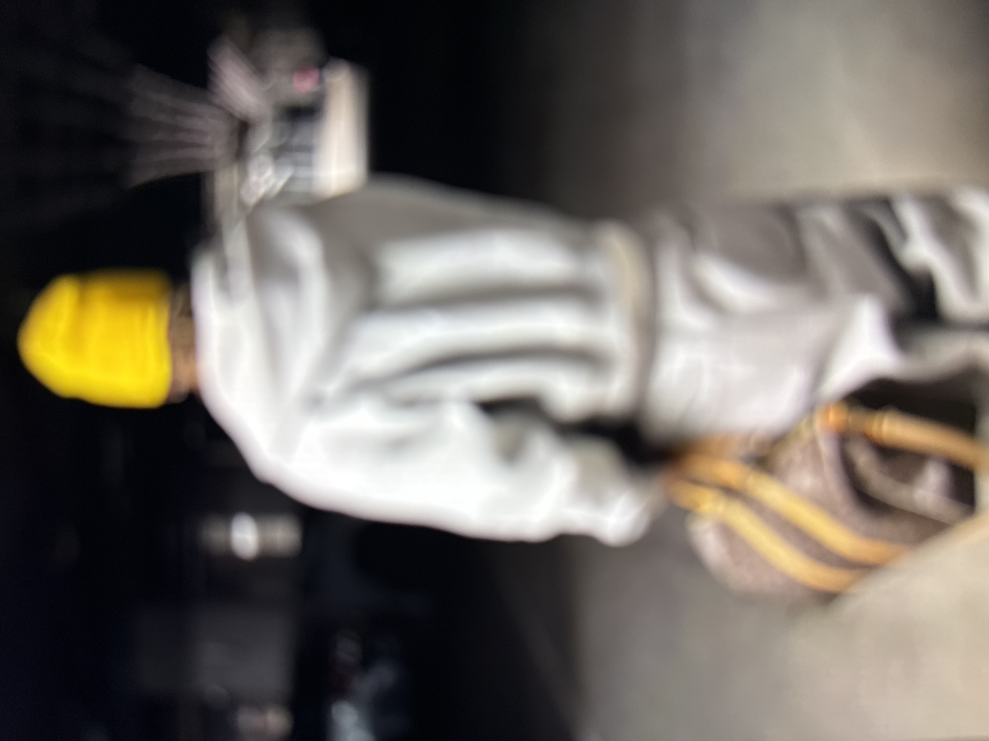 The image, captured with significant motion blur, depicts an individual from a rear perspective, walking away from the viewer in a dimly lit environment. The person wears a bright yellow beanie or cap, a light-colored (likely white or light grey) hooded sweatshirt, and light-colored pants. They are carrying a patterned duffel bag in their right hand, which hangs by their side. The bag features a dark pattern, possibly a monogram, with prominent tan or light brown straps and trim. The ground surface appears to be a uniform, light grey material, likely concrete. The background is dark and out of focus, showing indistinct blurred light sources and shapes, suggesting an indoor parking structure, a tunnel, or a similar urban setting at night. No specific architectural details or geographical features are discernible to identify the location as Kardzhali, Bulgaria, or any other specific city. No interactions with other people or subjects are visible.