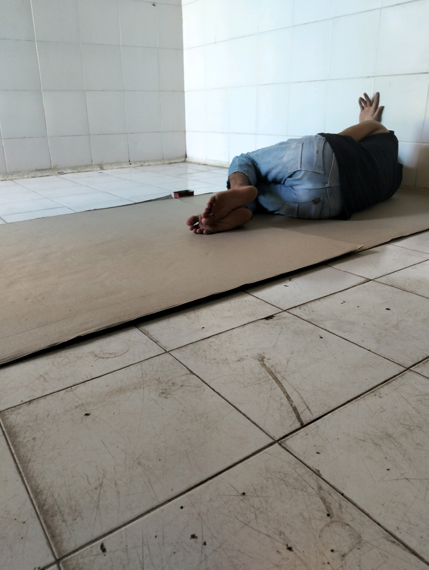 In a sparsely furnished room with white tiled walls and floors, a person is lying on their side on a large piece of cardboard, resting their head on their arms which are crossed behind it, leaning against the wall. They are wearing blue jeans and a dark t-shirt. The floor tiles are dirty and scratched. The room appears to be in Tanggulangin, Indonesia.