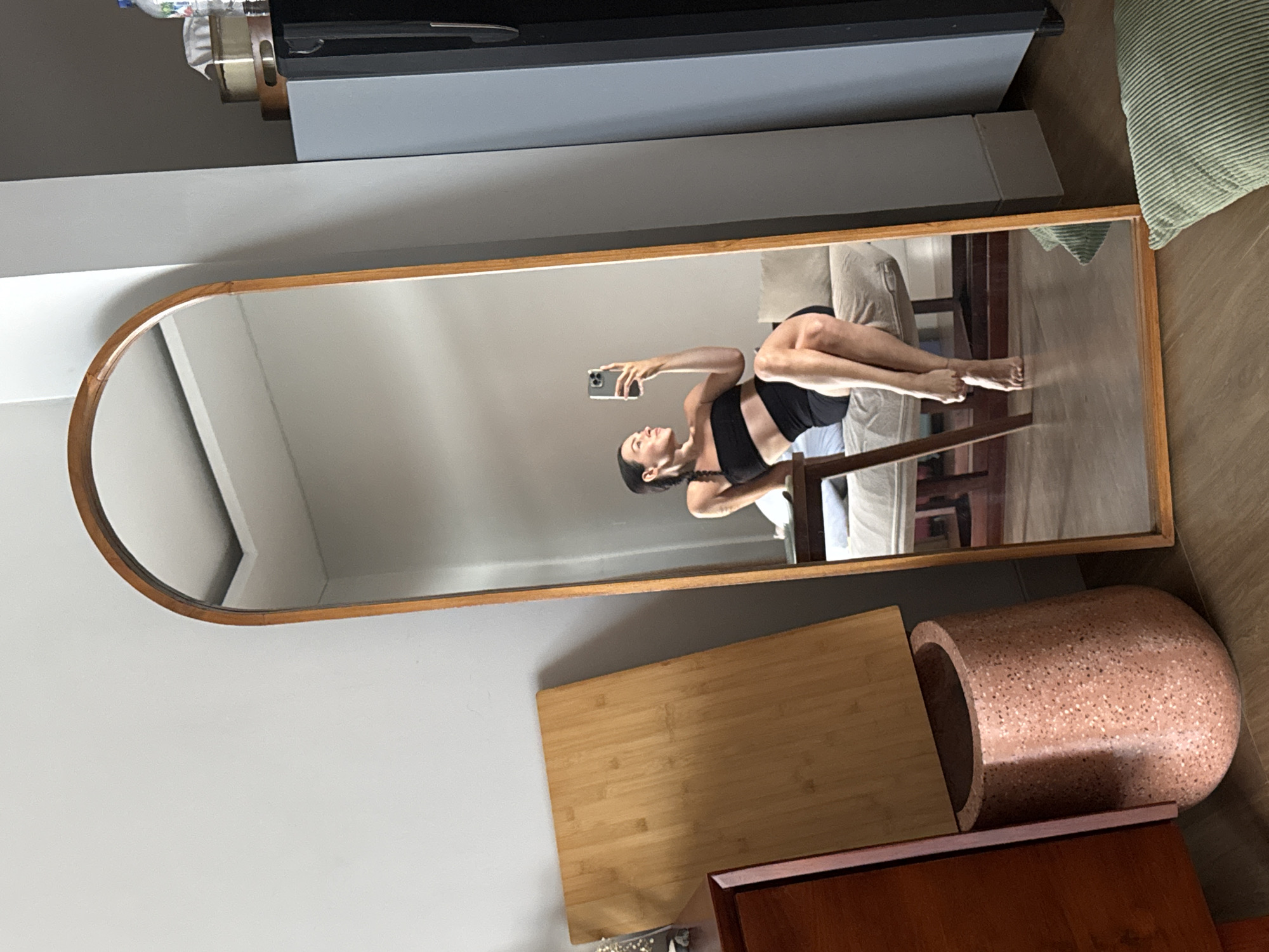 A woman is taking a mirror selfie in a room. She is sitting on a couch and wearing a black tube top and black shorts. She is looking at her phone and smiling. The mirror is large and oval-shaped with a wooden frame. In the background, there is a wooden table, a gray couch, and a gray wall. There is also a large wooden cutting board leaning against a dresser, and a pink speckled planter. The floor is made of wood. The image was taken in North Kuta, Indonesia.