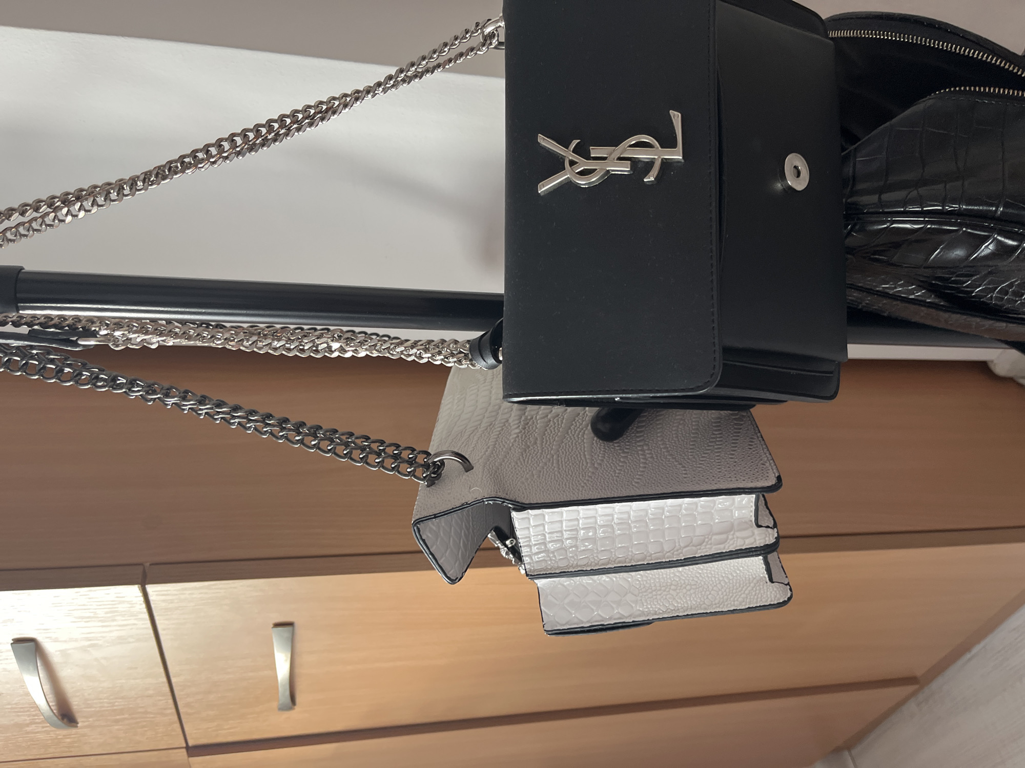 The image shows a black Yves Saint Laurent purse hanging from a black metal bar. A silver chain is attached to the purse, and the YSL logo is prominently displayed on the front. Below the black purse is a white and gray purse with a textured pattern, also hanging from the same bar by a silver chain. In the background, there is a wooden cabinet with silver handles. The environment appears to be an indoor closet or storage space. The location is unspecified and there is nothing to suggest it is located in Blagoevgrad, Bulgaria.