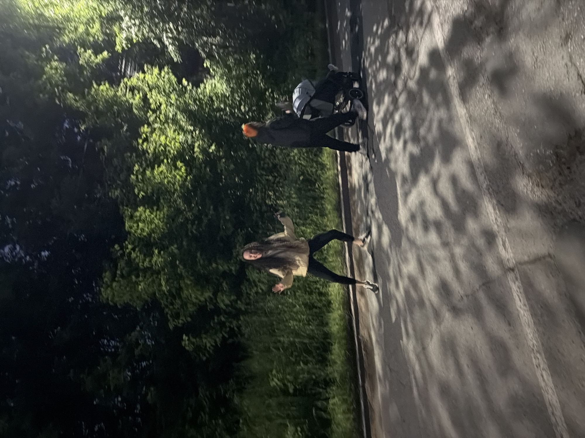 This nighttime photograph, taken in the city of Chaikovsky, Russia, depicts two individuals on a paved surface with dense foliage in the background. On the left, a woman with long dark hair, wearing a light-colored jacket, dark leggings, and sneakers, stands in a dynamic, mid-action pose with her arms raised, hands open, and a visible smile. To her right, an individual wearing a dark jacket, dark pants, and light-colored sneakers, characterized by bright orange hair or headwear, pushes a dark-colored stroller containing an occupant. This individual is seen in profile, moving from right to left across the frame. The paved ground exhibits distinct dappled shadows, indicative of tree canopies and artificial illumination, and a faint white line is visible horizontally on the asphalt. The background is composed of dark, abundant trees and undergrowth, with portions of the foliage on the right side brightly illuminated by an unseen light source, likely a streetlamp.
