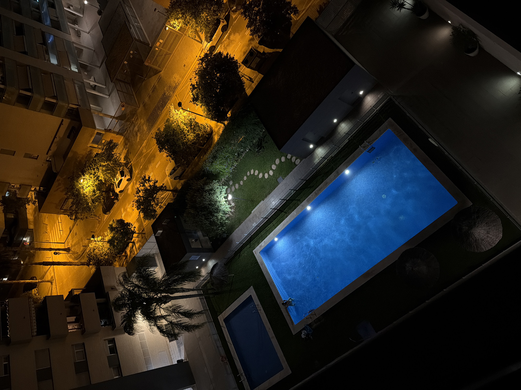 An overhead, night-time shot captures a view of two outdoor swimming pools, one smaller and rectangular, the other larger and rectangular, set within a grassy area. Both pools are illuminated by underwater lights, casting a bright blue glow. In the larger pool, a person is visible in the shallow end. Scattered around the pools are two thatched umbrellas, and a palm tree stands near the smaller pool. Beyond the grassy area, a paved pathway with stepping stones winds through lush greenery. A low fence separates the pools and garden from a street lined with trees and illuminated by streetlights. Several cars are parked along the street, and buildings with lit windows rise in the background. The overall scene is one of a residential urban environment at night.