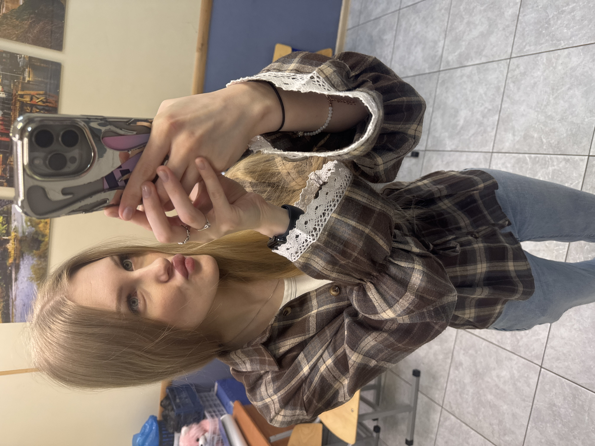 A young woman with blonde hair takes a selfie with a silver iPhone in an indoor environment. She wears a brown plaid shirt with white lace detailing around the sleeves. She is wearing blue jeans. The floor is tiled in a light gray. A cluttered area with what appears to be a blue storage container and other objects is partially visible. Wall art hangs on the wall. The location may be Perm, Russia.