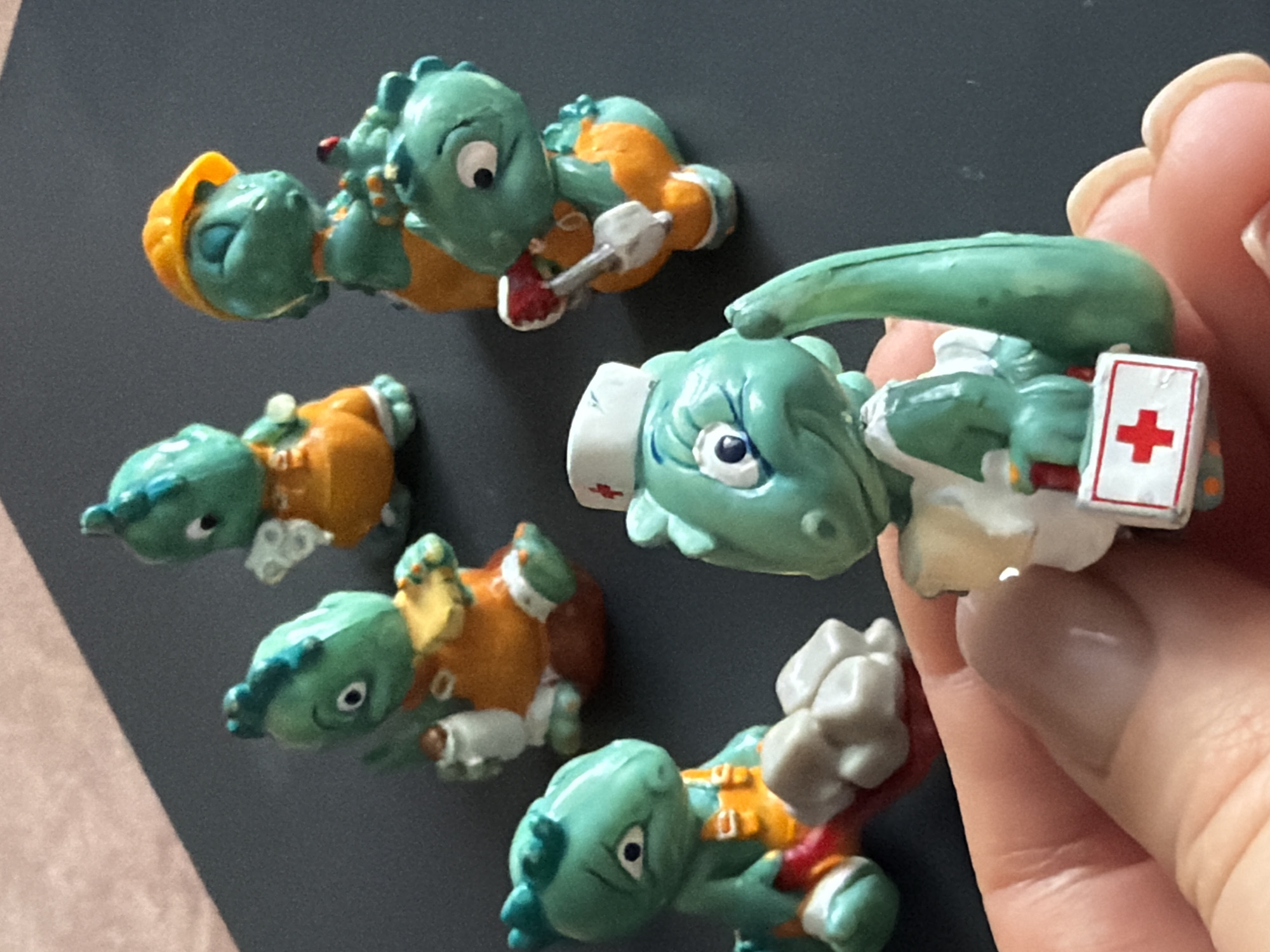 The image shows five small, green, cartoon dinosaur figurines. Each dinosaur is dressed as a different professional, including a construction worker with a yellow hard hat, a dentist with a toothbrush, a nurse with a white nurse's cap and first aid kit, and two others wearing orange vests. The figurines are placed on a dark gray surface, and a hand is holding one of the figures (the nurse). The location is not identifiable as Krasnoyarsk, Russia, or any other specific city based on the image.