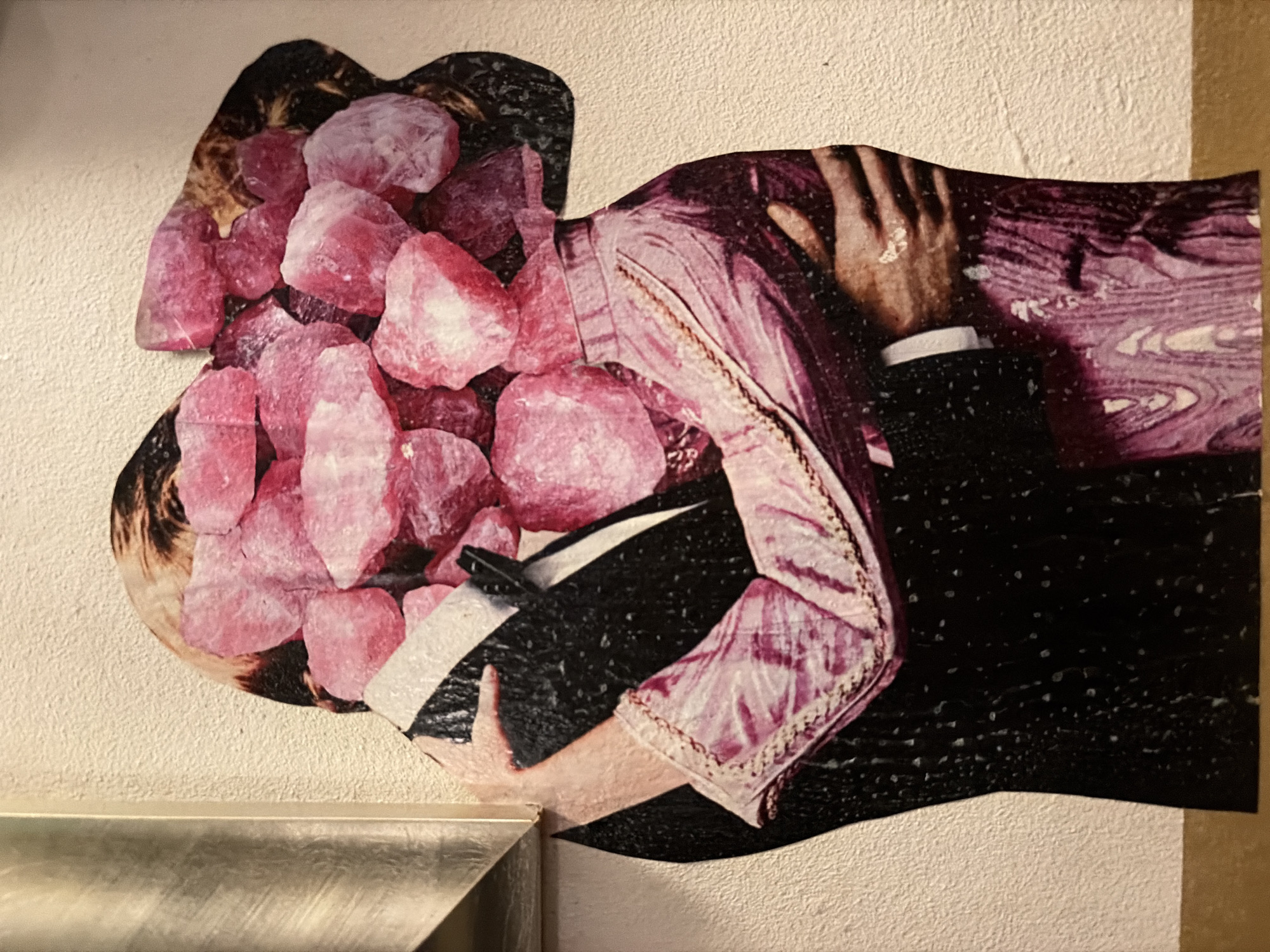 The image depicts a collage of two figures embracing. The face of the first figure is composed of raw pink crystals. The second figure is wearing a black suit and a pink and white patterned garment. A hand with a ring is visible. The collage is adhered to a white textured wall above a silver-colored surface, presumably a ledge or countertop. There is no evidence in the image to suggest this image is located in Barcelona, Spain.