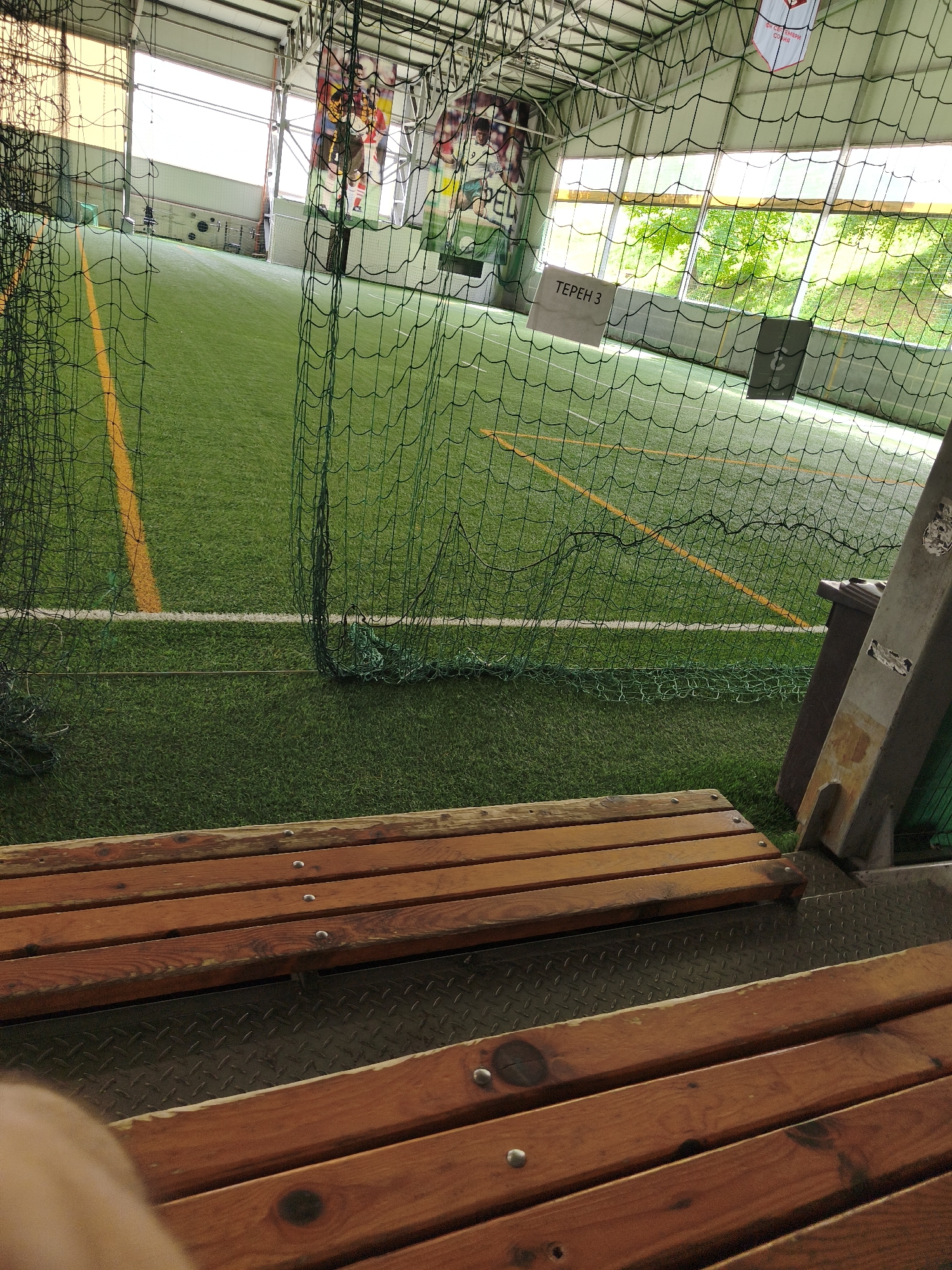The image shows an indoor football field. The field has artificial turf with yellow and white markings. A green net encloses the field. There are benches in the foreground, made of wood with metal fasteners. In the background, there are large windows offering a view of green trees and foliage. There are also banners hanging from the ceiling with images and text, one of which reads "ТЕРЕН 3" (Field 3) and another appears to be a sports club banner with Cyrillic text. The overall impression is of a sports facility.