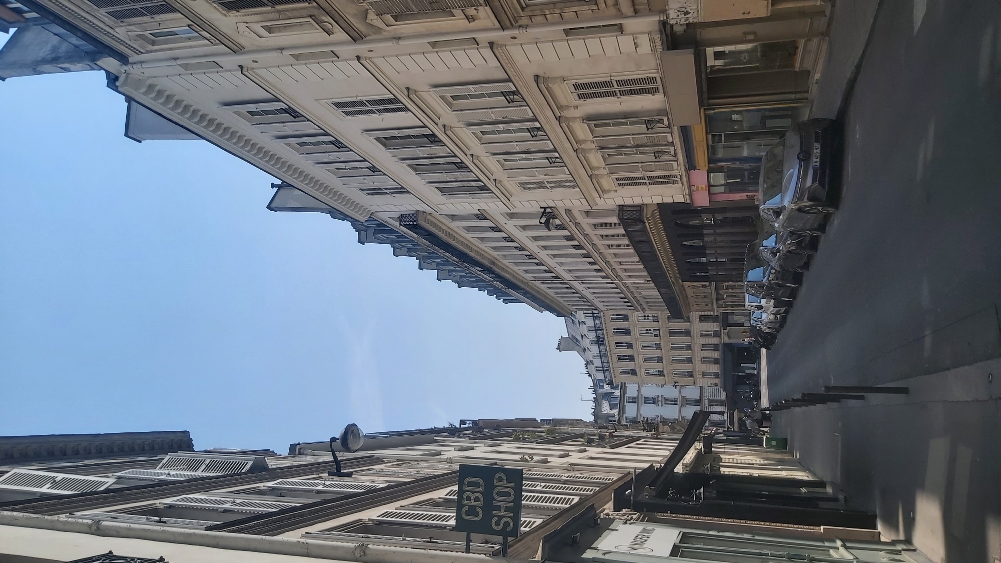 The image depicts a street scene in Paris, France, with classic Haussmannian buildings lining both sides of a narrow road. The architecture is characterized by pale facades, mansard roofs, and numerous windows with shutters, indicative of a traditional Parisian urban landscape. On the left side of the street, a sign reads "CBD SHOP" in green lettering, suggesting a commercial establishment. Several parked cars are visible along the right side of the road, suggesting a residential or mixed-use area. The sky is clear and blue, indicating fair weather. The perspective is from street level, looking down the length of the street.