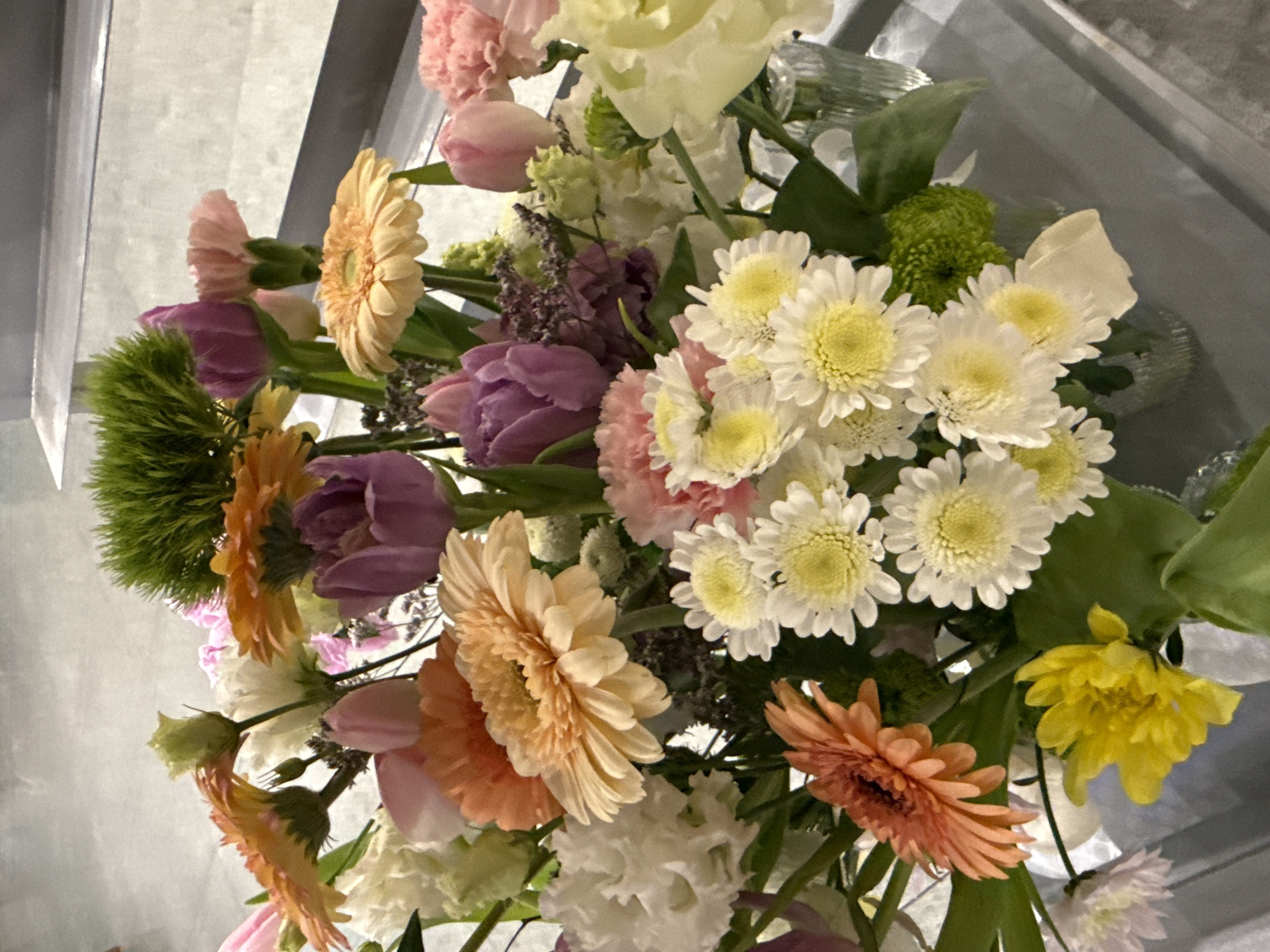 A close-up, slightly tilted shot displays an abundant bouquet of mixed flowers. Gerbera daisies in shades of orange and peach are prominent, interspersed with white daisies, purple tulips, pink carnations, and a green globular bloom. The arrangement also includes yellow flowers and sprigs of greenery. The background is a blurred gray, giving no indication of location. The absence of identifiable landmarks or cultural indicators prevents determining if the image was taken in Saint Petersburg, Russia.