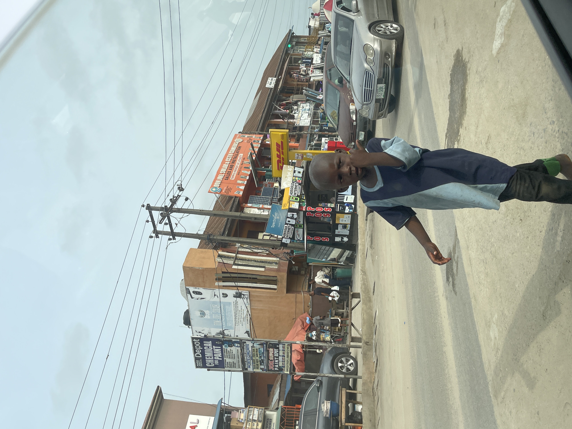 This candid daytime photograph captures a street scene in Ado, Nigeria, likely taken from inside a moving vehicle due to the visible reflection. The main subject is a young boy in the foreground, wearing a blue and light blue long-sleeved tunic over dark trousers, with a green patch visible on his left foot. He looks directly at the camera, his right hand slightly raised and open, with a neutral to slight smile expression.
Behind the boy, the image reveals a busy commercial street with several buildings housing various businesses. Utility poles with a dense network of overhead wires line the street. Cars are present, including a silver Mercedes-Benz sedan partially visible on the right with the license plate "FST 32DL," and a darker vehicle further down the street. Other individuals can be seen in the background near the shops.
Numerous signs for local businesses and services are prominently displayed. Visible text includes "DON JOEL SOLAR & CCTV CAMERAS SALES AND INSTALLATION," "MAMA Depot CHEMICAL FOR PAINT," "MAMA JOHNTIT CHEMICAL INCRETE CONCRETE," and "JOK SUPER PAINT DEPOT." Other signs advertise "airtel 4G," "ADEGOLD INTERNET," "Spectranet," and "POS." A DHL logo is also visible. Solar panels are displayed outside the "Don Joel Solar" establishment. The sky is overcast, indicating a cloudy day, and the street appears to be asphalt or concrete with some stains and markings. The overall atmosphere suggests an active urban commercial area.