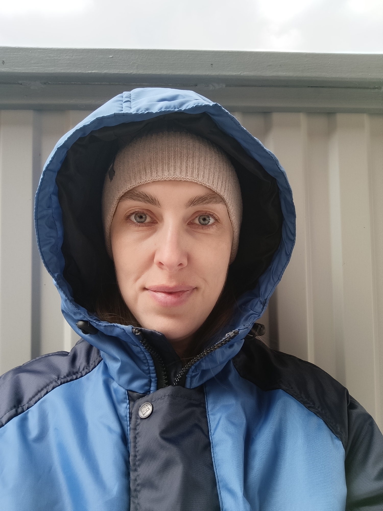 A close-up portrait features a woman posing outdoors, seen from the chest up against a plain background. She has light-colored eyes, a subtle smile, and is looking directly at the camera. She wears a light beige knitted beanie and a two-toned winter jacket. The jacket is primarily light blue on the main body and shoulders, with dark blue or black sections on the lower sleeves and accents. The hood is pulled up, framing her face, and is also dark blue on the inside lining. The jacket has a black zipper and a prominent silver snap button on the front with a discernible, stylized 'B' or similar letter engraved within a circle.
The background consists of a light grey or off-white corrugated metal wall or paneling, suggesting an industrial or utilitarian structure. Above this wall, a thick horizontal grey beam or ledge is visible, with a cloudy, overcast sky above it. The lighting is diffused, indicating a daytime setting under cloudy weather, consistent with the warm attire. The overall mood is calm and neutral. The setting is likely an outdoor, possibly urban or industrial, location in Chaikovsky, Russia.