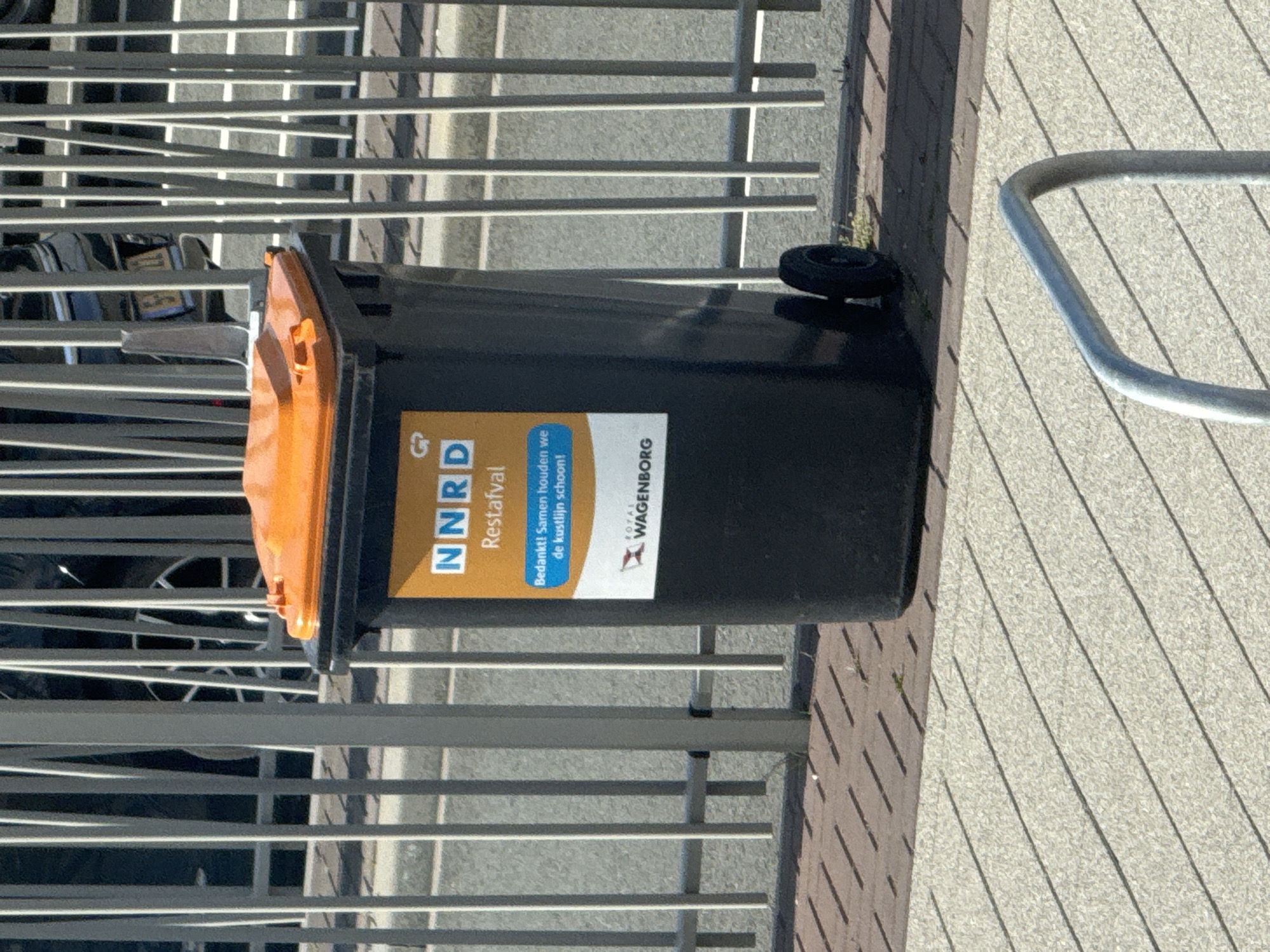 A black waste bin with an orange lid, equipped with two black wheels, stands outdoors. A yellow and blue label on the bin prominently displays "NNRD Restafval" (Residual Waste), a blue band with "Bedankt! Samen houden we de kustlijn schoon!" (Thanks! Together we keep the coastline clean!), and a "ROYAL WAGENBORG" logo at the bottom. The bin is positioned in front of a grey metal fence composed of vertical bars. Behind the fence, a paved road or parking area is visible, with parts of two dark-colored vehicles obscured; one on the left, and another on the right revealing a partial yellow license plate. The ground consists of reddish-brown diagonal brick paving directly beneath and behind the bin, transitioning to a lighter grey, speckled surface with parallel diagonal lines in the foreground. A silver, curved metal rail, consistent with a bicycle rack, is partially visible in the bottom center foreground. The scene is illuminated by strong daylight, casting distinct shadows. The location is in the city of Unknown, Unknown.