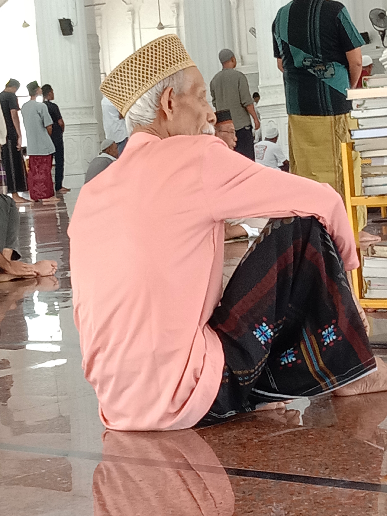 An elderly Muslim man, wearing a peach-colored robe and a woven tan prayer cap, is seated on a polished floor. He is looking away from the camera and his left hand rests on his lap. He is wearing a black sarong with red, blue, and white geometric patterns. In the background, other people are visible, some standing and some seated. One person in the background is wearing a black and green striped shirt and a tan sarong. Another person is wearing a gray shirt and dark pants, and a third person is wearing a black t-shirt and dark pants. The location appears to be inside a building with white pillars and a tiled floor. The image may have been taken in Lhokseumawe, Indonesia.