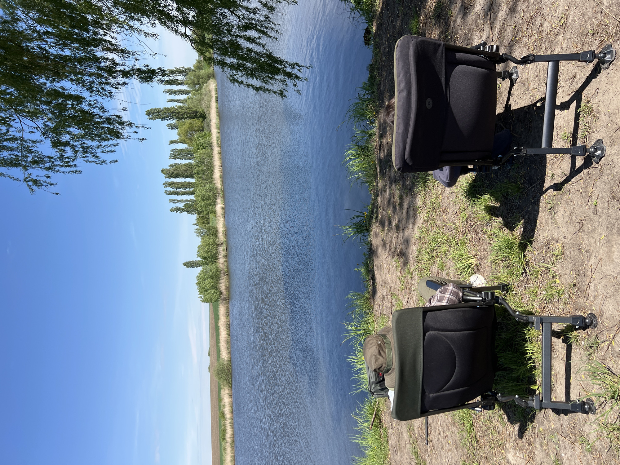 The image shows a waterside scene in Romanove Selo, Ukraine. Two black fishing chairs are placed on a grassy bank, adjacent to a wide body of water, likely a river or canal. The water extends horizontally across the majority of the frame. On the opposite bank, a row of trees and vegetation are visible. The sky above is clear and blue, with some trees branches partially obscuring the sky at the top left.