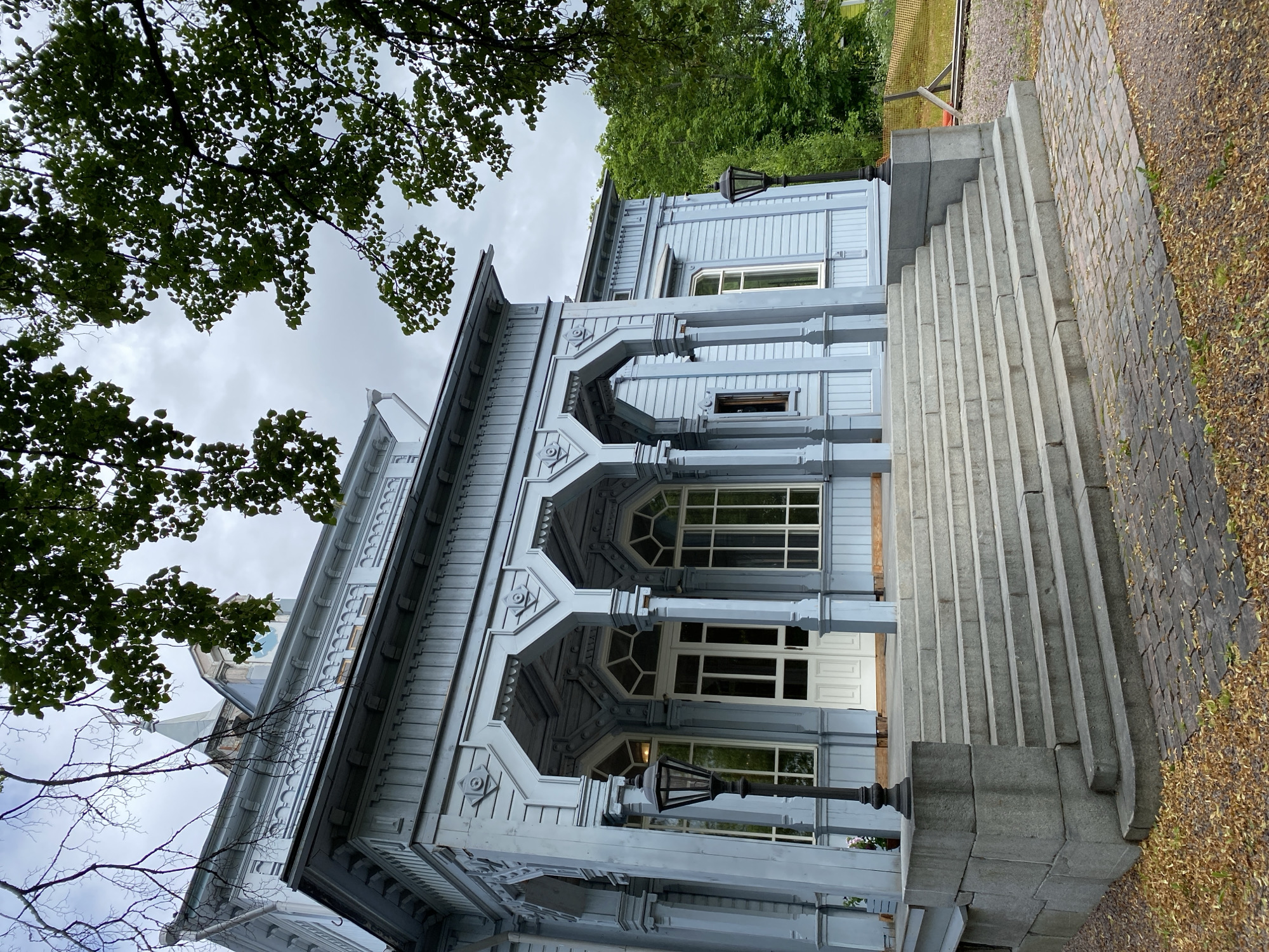 The image depicts the exterior of a light blue wooden building, likely a historic house or museum, in Sortavala, Russia. The building features intricate architectural details, including decorative trim, arched verandas with columns, and large windows. A wide, stone staircase with multiple steps leads up to the main entrance, which is marked by a white double door. Two black lampposts stand on either side of the staircase. The building is surrounded by greenery, with trees and foliage visible in the foreground and background. The sky is overcast, suggesting a cloudy day. The overall impression is one of old-world charm and historical significance.