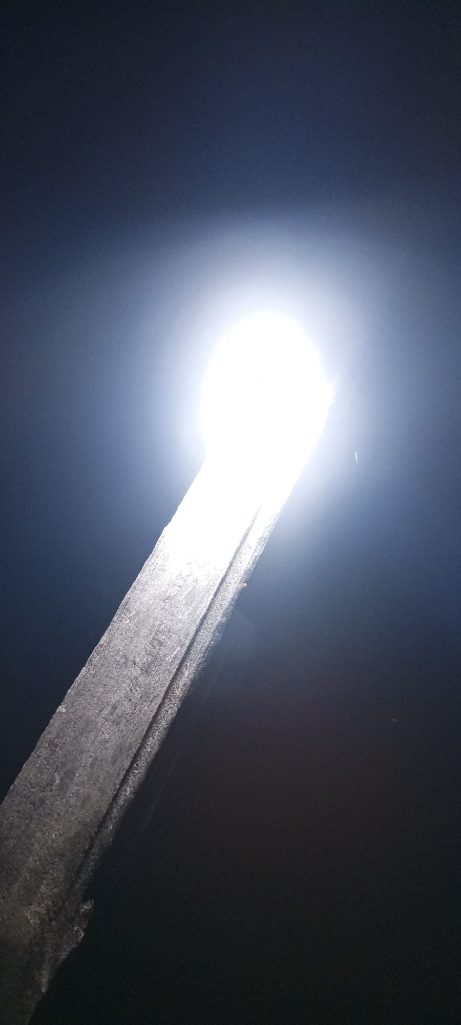 The image depicts a brightly illuminated wooden beam extending diagonally upwards from the lower left corner against a dark background, possibly at night. The source of the light is not clearly visible but appears as a bright, circular, almost blinding orb at the top of the beam. The setting suggests an outdoor location. The specifics to the city of Adankolo, Nigeria, are not discernible from the visual information provided.