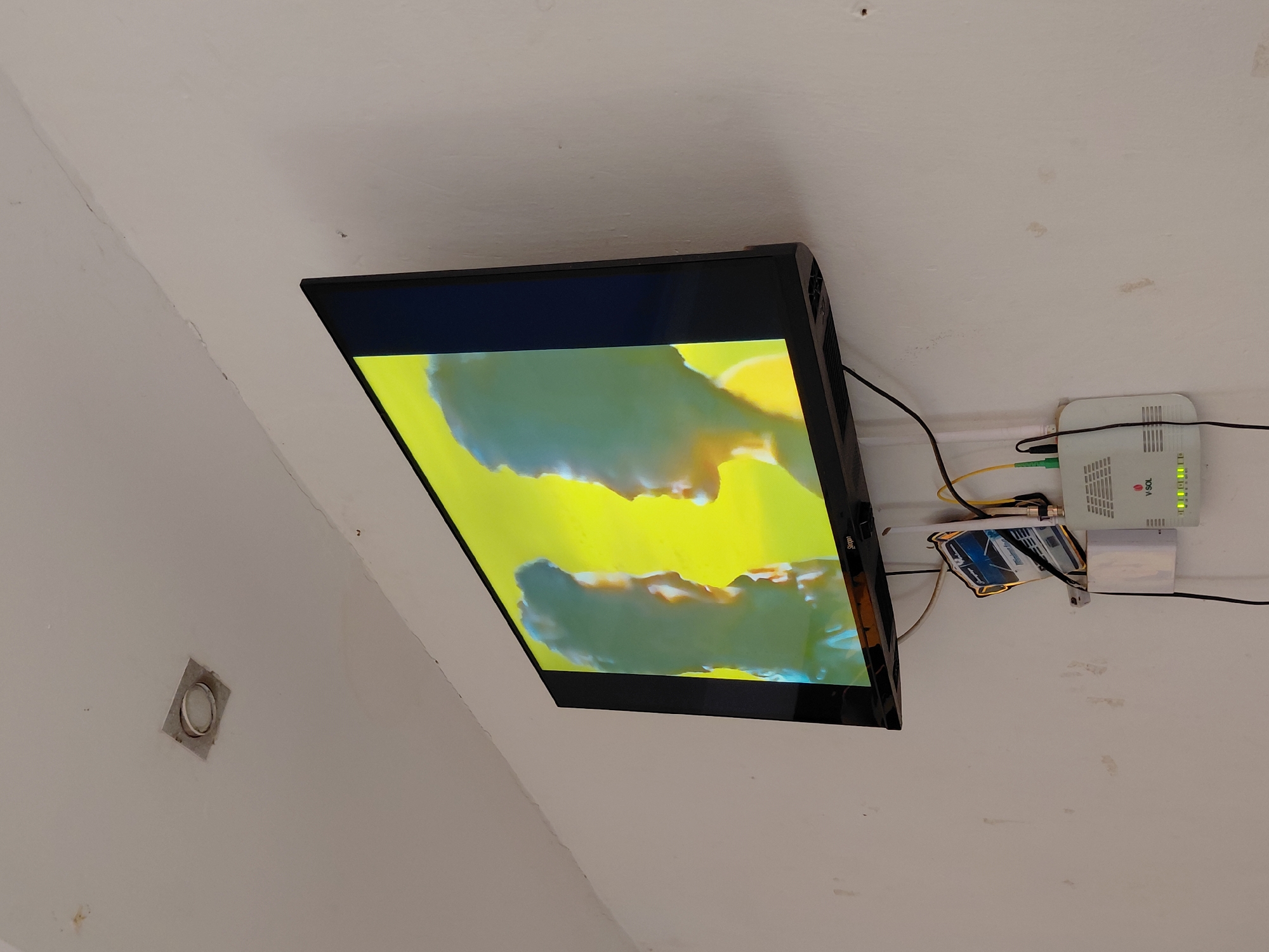 A flat-screen television is prominently displayed, mounted on a textured white wall, and is actively displaying a vibrant image characterized by bright yellow and blue-green hues. The TV's frame is black, and the visible brand name on its side is "Sugor."
To the right of the television, a white internet router, branded "V-SOL" and labeled as a "Fiber Optic Terminal," is affixed to the wall. Its green indicator lights are illuminated, suggesting it is powered on and operational, with "PON," "LAN," "TEL," and "POWER" lights visible. A cluster of cables, including yellow, green, and black wires, connects to the router and extends around the setup.
Below the router, a small, rectangular object with a glossy finish is mounted, featuring a picture of a car and a discernible "Lamborghini" logo. The white wall itself shows signs of wear, including faint cracks, smudges, and areas of discoloration, particularly in the upper left corner and near the router. A small, square-shaped vent or outlet with a circular white insert is visible on the wall in the lower-left section of the image. The overall setting appears to be an indoor space, possibly a room, with functional electronic equipment mounted directly onto the wall.