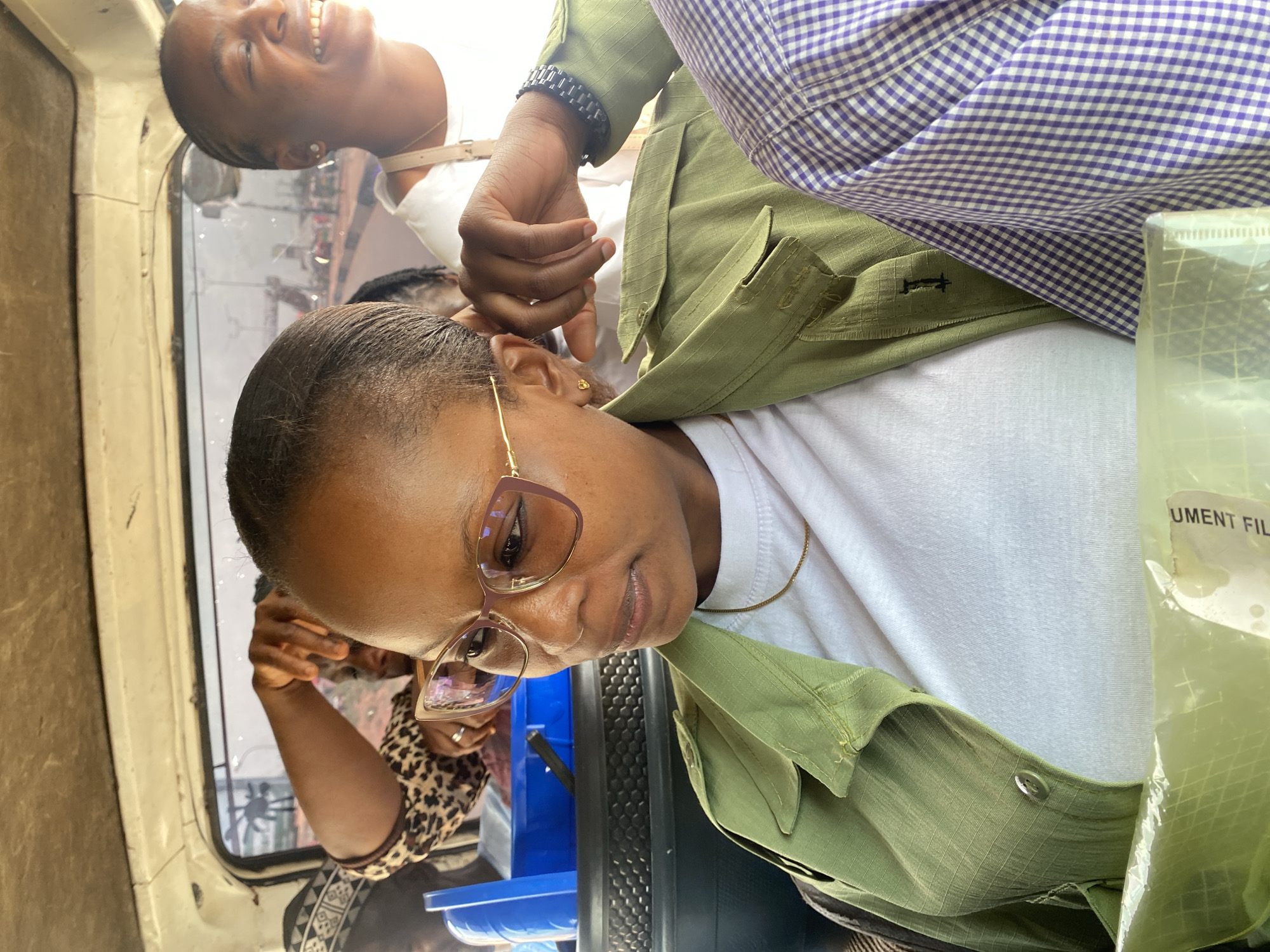 The image captures a scene inside a vehicle in Gura Top, Nigeria. A woman wearing glasses, a white t-shirt, and an olive green jacket is the primary subject. A hand with a dark-colored bracelet is reaching towards her ear. Above and behind her, another woman is visible, smiling and looking out the window. Other passengers are partially visible in the background. The interior of the vehicle appears to be a public transport vehicle, with a window and other details suggesting a shared space. A plastic bag is in the foreground. The overall setting indicates a typical urban transport environment in Gura Top.