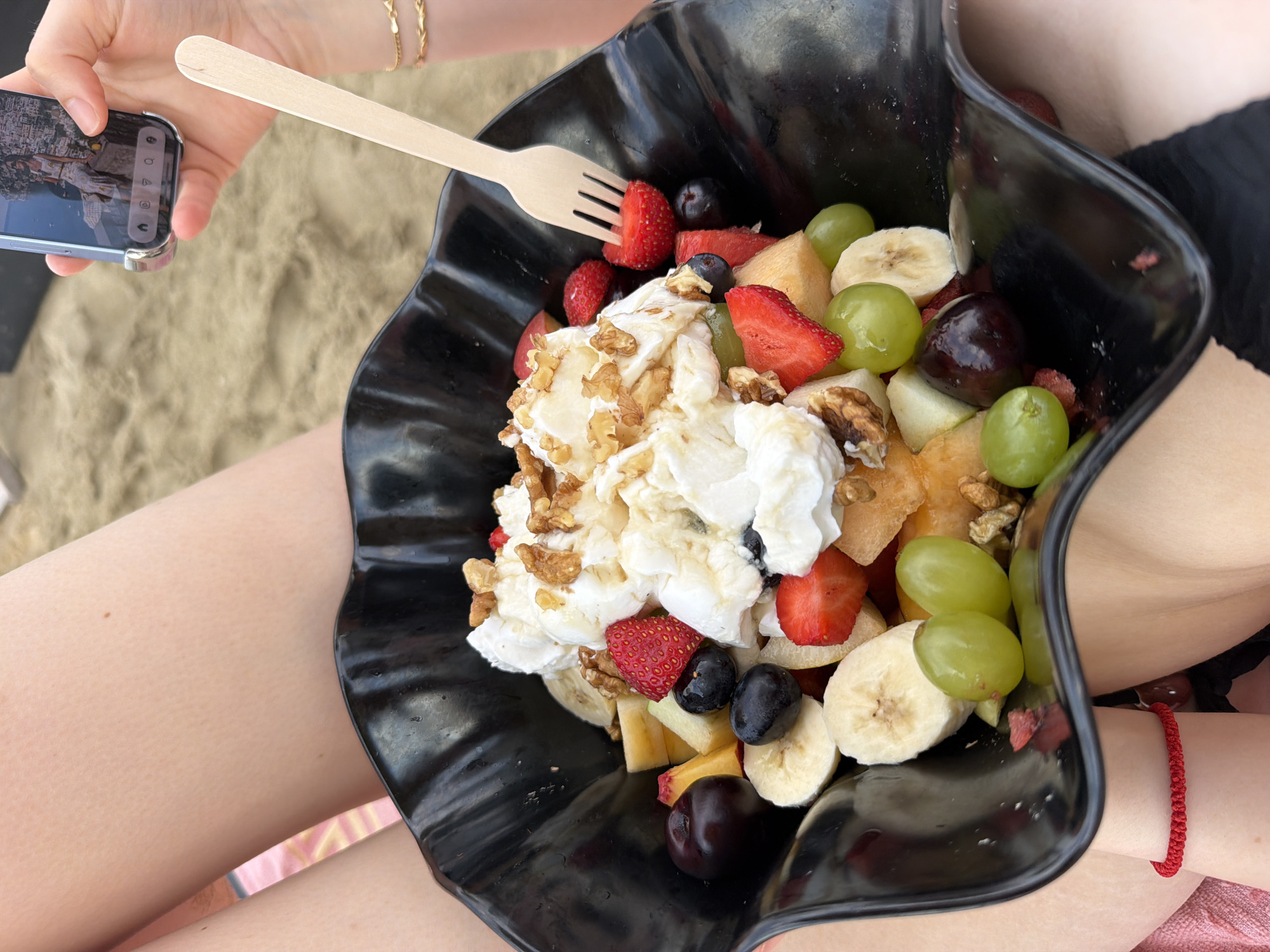 A close-up overhead shot shows a black, wavy-edged bowl filled with a fruit salad on a sandy beach, likely in Stavros, Greece. The fruit salad contains strawberries, blueberries, grapes, bananas, cherries, and possibly melon, topped with whipped cream and walnuts. A light-colored wooden fork is in the bowl, piercing a strawberry. In the upper right corner, a person's hand holds a smartphone, displaying a photo. Another person's legs, wearing a red string bracelet, are visible in the foreground and on the left. The background is out of focus, revealing more sand and hints of beach attire.