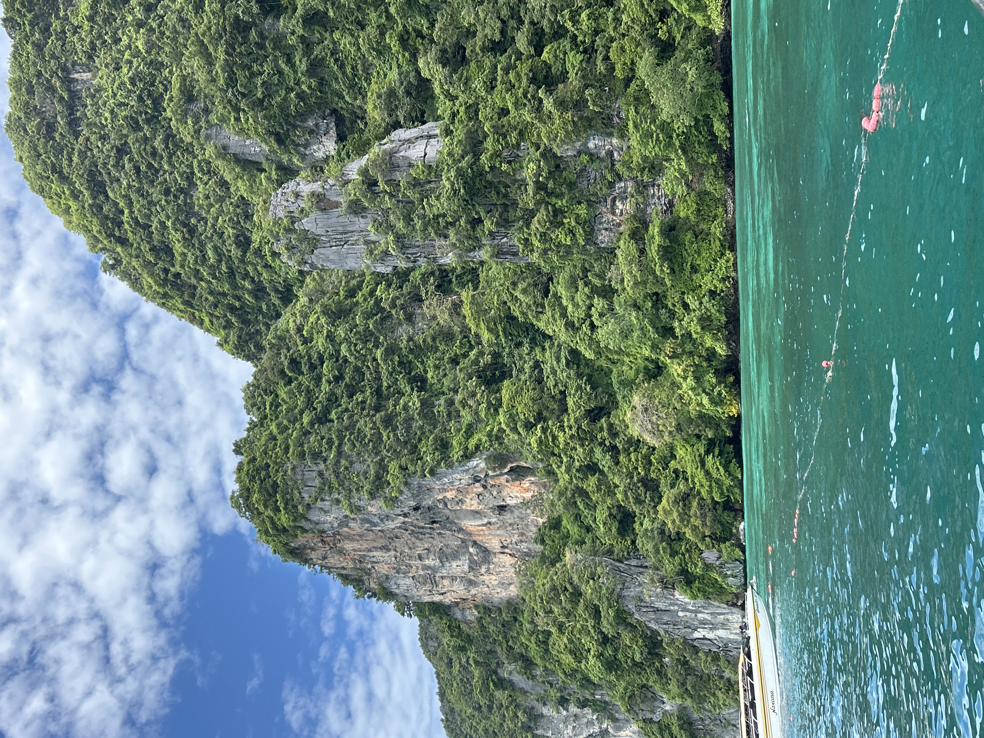 The image depicts a scenic view of a tropical coastline, likely near Ao Nang, Thailand. In the foreground, clear turquoise water stretches towards a lush, green, mountainous island. The island is characterized by steep, rocky cliffs covered in dense vegetation. A long-tail boat, a common type of vessel in Thailand, is visible on the left side of the image, its white and yellow hull partially submerged in the water. The boat appears to be moving or anchored near the shore. Several pink buoys are scattered across the water, connected by a rope, suggesting a designated swimming or boating area. The sky above is a vibrant blue, dotted with fluffy white clouds. The overall scene evokes a sense of natural beauty and tropical leisure.