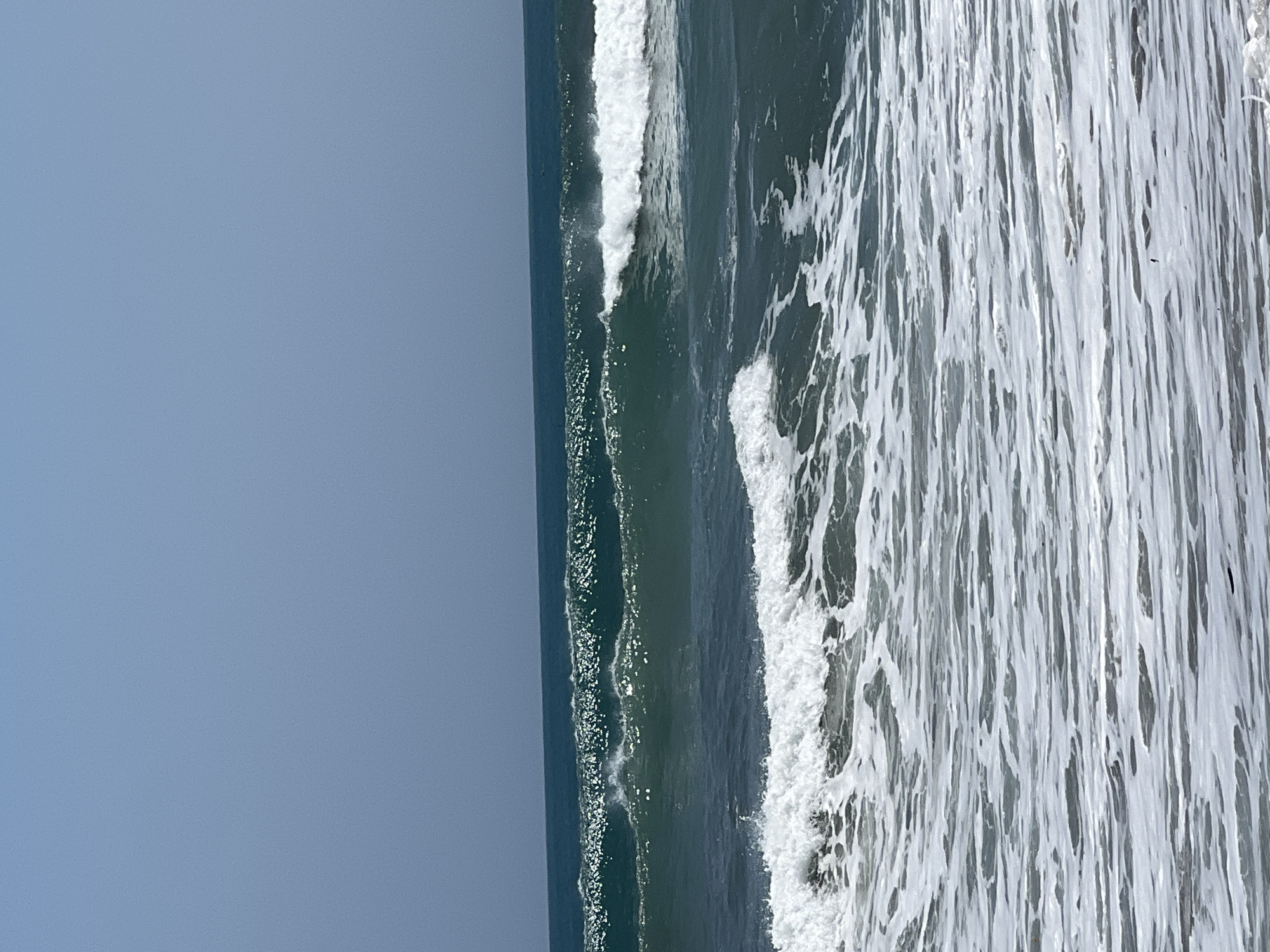 The image shows a view of the ocean with waves breaking. In the foreground, there are foamy white waves rolling in. Further out, there are larger, darker green waves, some with white crests. The sky above is a clear, pale blue. The scene depicts a typical beach environment, with no people or structures visible. The location is identified as Kuta, Indonesia.