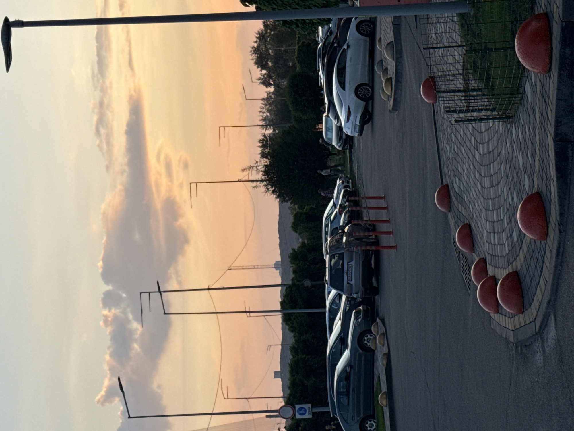 The image captures an outdoor scene at what appears to be sunset or dusk, characterized by a sky that transitions from light yellow to soft orange and pink, with scattered, warm-toned clouds.
In the foreground and midground, a line of cars is parked along a paved road or parking lot. The vehicles are various models and colors, predominantly white, silver, and dark grey. Adjacent to the road, a pathway or sidewalk is visible, featuring a distinct pattern of interlocking brick pavers. Along this path, several large, red, half-dome shaped barriers are placed at regular intervals. A low metal fence separates the paved path from a grassy area.
Numerous tall streetlights with horizontal arms extend into the sky, their dark silhouettes contrasting against the bright horizon. Beyond the parking area, a dense line of green trees forms a natural barrier, with a hazy, darker landscape, possibly distant hills or urban structures, visible in the background.
A few individuals are present in the scene; one person appears to be walking near the parked cars, and others are partially visible further back among the trees. Near the road, a blue rectangular sign with white text is clearly visible, displaying "ЗОНА" (ZONA, meaning "zone") and "30м" (30 meters). Mounted above this, on the same pole, is a circular sign with a red border and a diagonal line, typically signifying "No Stopping" or "No Parking." The overall impression is one of a calm, public outdoor space during the late hours of the day.