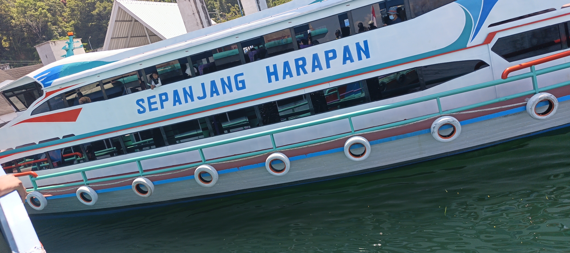 A side view of a white passenger ferry named "SEPANJANG HARAPAN" is shown partially submerged in green water, angled from lower left to upper right. The ferry has multiple windows along its side, through which passengers are visible. The hull of the ferry is light gray with a maroon stripe and a blue stripe above it. A turquoise railing runs along the deck, with red accents. Several white tires with orange centers are attached to the side of the hull. In the background, a building with a white roof and a gray facade is visible, with trees and foliage beyond it. The scene appears to be in a harbor or bay.