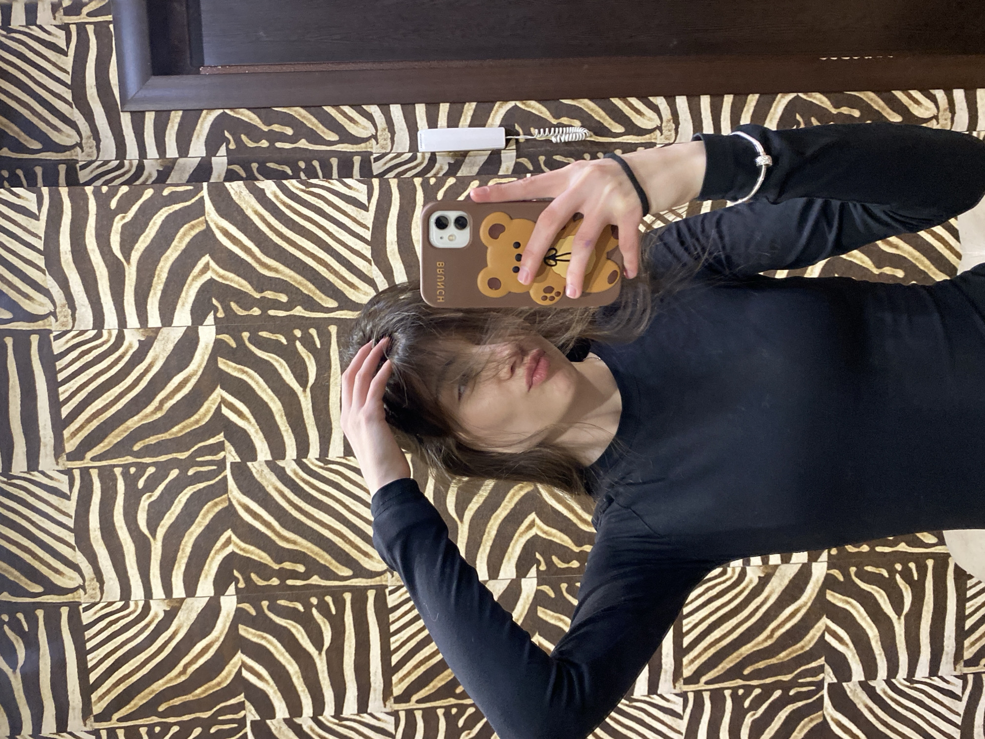 A young woman is taking a selfie in front of a zebra-striped wall or surface. She is wearing a black long-sleeved shirt and has a phone case with a brown bear design. The location is Votkinsk, Russia. There is a white object and a small brush on the wall above her. The photo is taken from a slightly elevated angle, capturing the woman's face and upper body, along with the patterned background.