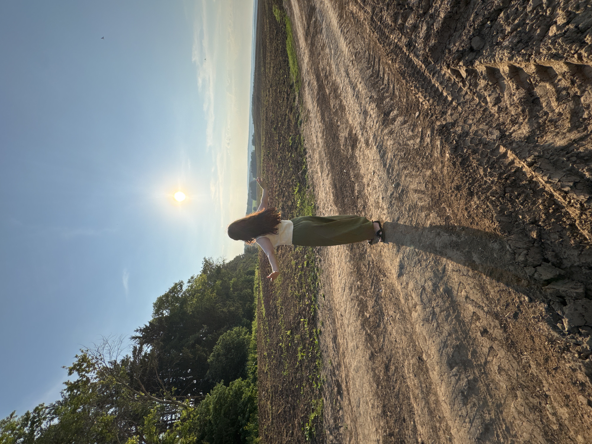 In Lukintsy, Russia, a young woman with long, dark hair stands in a field with her back to the camera. She is wearing a white top and a long, olive-green skirt, and her arms are outstretched to her sides. The sun is shining brightly in the sky, casting a long shadow behind her. The ground is a dirt path with tire tracks, and the field is partially tilled with some patches of green grass. In the background, there are trees and rolling hills. The sky is clear blue with a few scattered clouds. There are also two small birds flying in the sky.