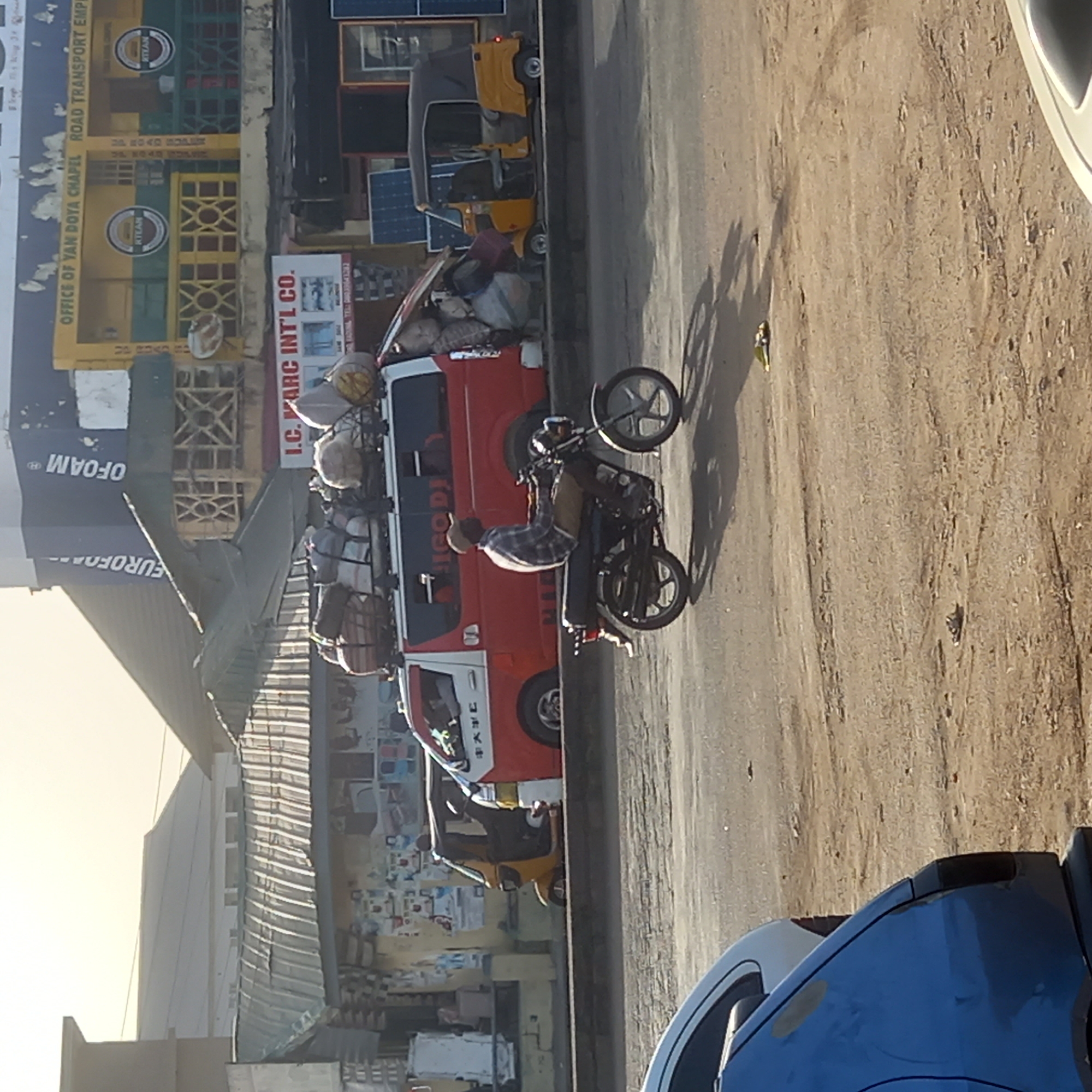 The image captures a vibrant street scene in what appears to be an urban or semi-urban setting, consistent with Dadi Riba, Nigeria. The time of day seems to be mid-day or afternoon, indicated by the bright sunlight and prominent shadows stretching across the dusty ground. The weather is clear and sunny.
In the foreground, a person wearing a plaid shirt and a light-colored cap is actively riding a black motorcycle. The motorcycle is depicted performing a wheelie or leaning significantly, with its front wheel raised off the ground, suggesting motion or an acrobatic maneuver. To the left of the motorcycle, a red and white truck or van is parked or moving slowly. This vehicle is heavily laden with numerous sacks and luggage piled high on its roof rack and in its open rear section. Another yellow tricycle, commonly known as a tuk-tuk or Keke Napep, is visible behind the red truck, and a similar one is further down the street. Part of a blue vehicle is visible in the lower right foreground.
The street itself features a paved section to the left and a wider, dusty, unpaved area to the right where the motorcycle is riding. Buildings line the street, showcasing various storefronts. A prominent yellow and green building displays "OFFICE OF YAN DOYA CHAPEL ROAD TRANSPORT EMPI" and two circular "KTEAN" logos. Below it, a red sign reads "I.C. MARC INT'L CO." Further left, a dark blue building or sign shows "EUROFOAM®" and "OFOAM®". Chinese characters "中大" are also visible on the side of the red truck. The overall scene conveys a busy commercial and transportation hub.