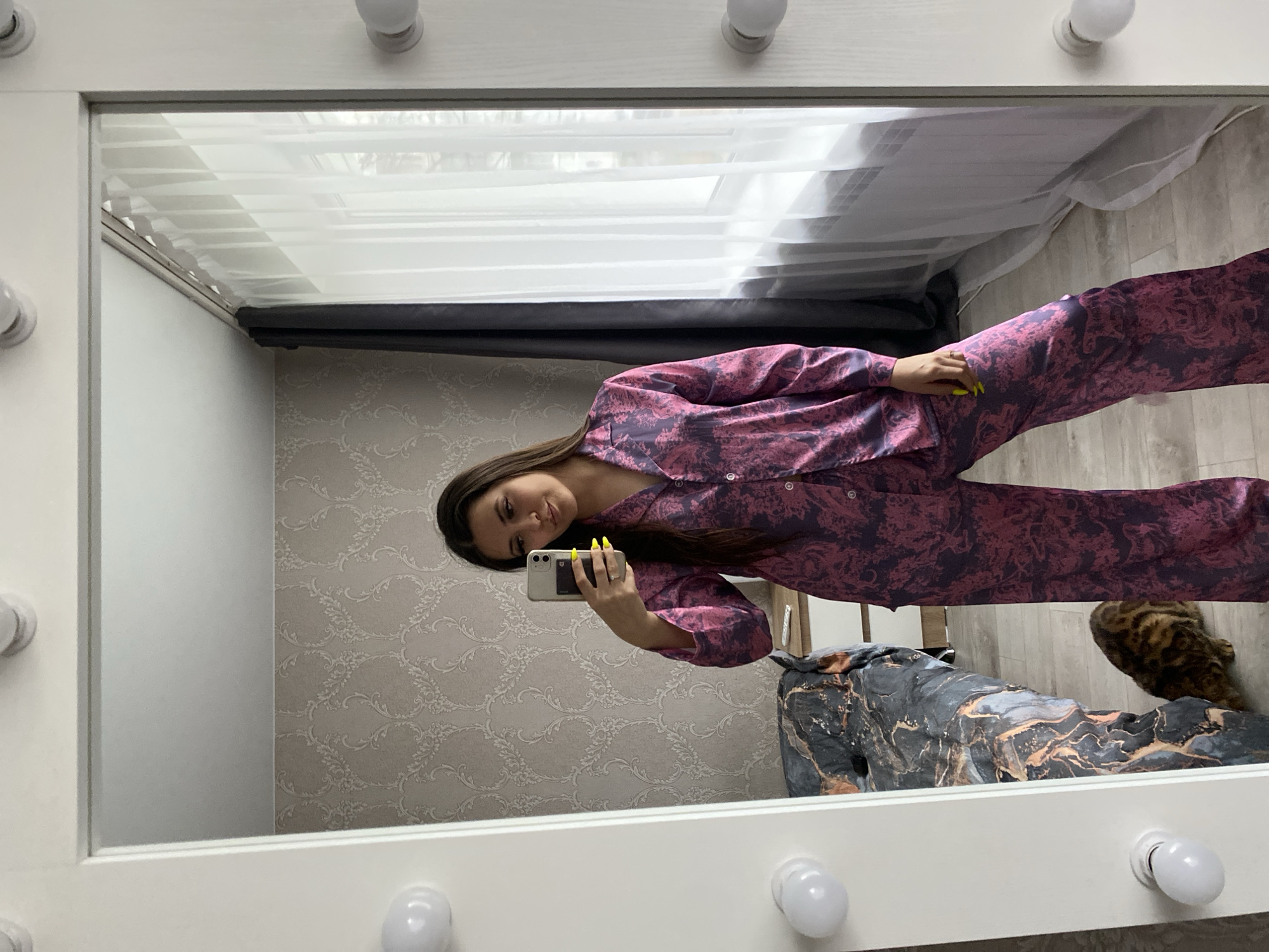 A young woman is reflected in a mirror taking a selfie in an indoor setting. She is wearing a matching pink and purple patterned pajama set. She holds a cell phone with bright yellow nail polish on the hand holding it. The mirror frame is white with decorative light bulbs. The background includes a wall with a neutral, patterned wallpaper, a window with sheer and dark-colored curtains, a bed covered with a marbled grey and gold blanket, and a spotted cat. It is unknown if this image was taken in Chaikovsky, Russia.