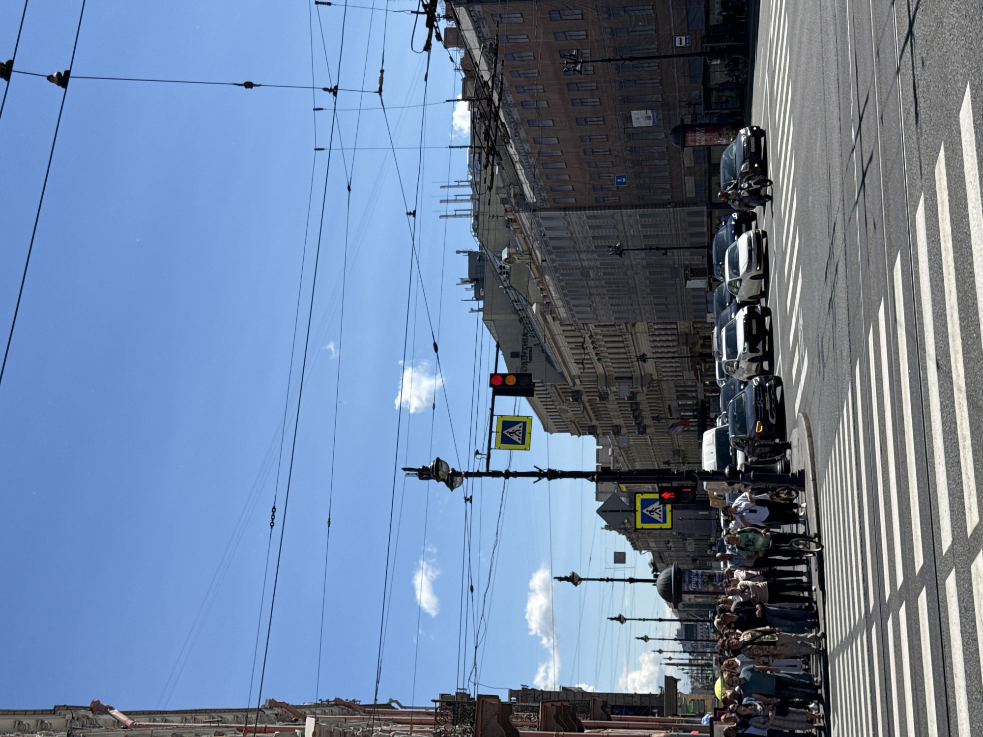 This is a photograph of a street scene in Saint Petersburg, Russia, on a clear, sunny day. The image captures a wide intersection with a pedestrian crossing in the foreground. Several cars, including a black Mercedes SUV, are visible on the road, along with a motorcycle. A group of pedestrians is waiting to cross the street, some looking towards the camera. Traffic lights and pedestrian crossing signs are present, with one traffic light showing red.
The architecture lining the street consists of multi-story buildings with classical facades, typical of Saint Petersburg. Overhead, a complex network of tram and trolleybus wires crisscrosses the sky, suggesting the presence of public transportation in the area. The sky is a bright blue with a few scattered white clouds. The overall impression is a bustling urban environment under a bright summer sky.
