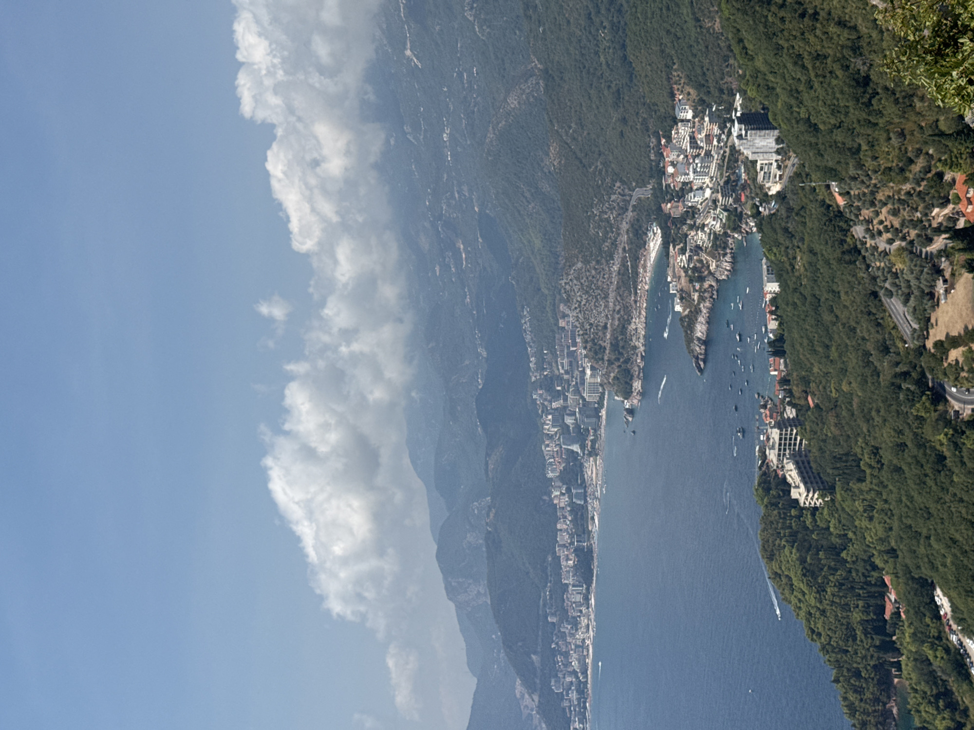 An aerial, full shot captures the coastline and mountainous terrain surrounding Sveti Stefan, Montenegro. The deep blue sea extends to the horizon, meeting a sky dotted with scattered white clouds. Along the shore, a densely packed town with numerous buildings, mostly white with red roofs, sprawls across the landscape. The coastline is characterized by rocky outcrops and a few small beaches. Further inland, lush green forests cover the steep, undulating hills and mountains. A winding road is visible snaking through the foliage, with parked cars near a building nestled in the trees. Boats are scattered in the water, suggesting some maritime activity. The scene is bathed in bright daylight, indicating a clear, sunny day.