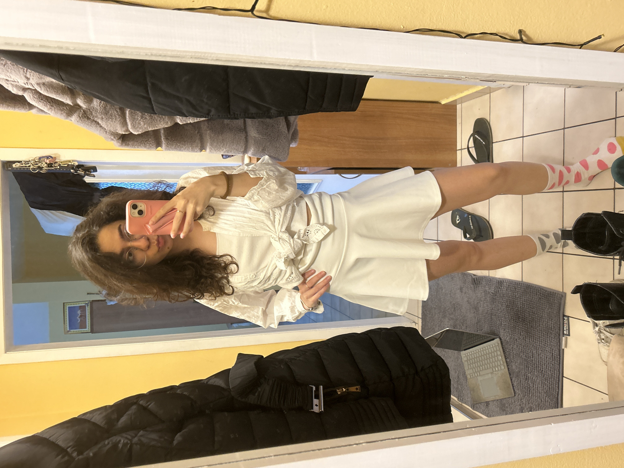 A person is taking a mirror selfie in a room. The person has dark curly hair, wears glasses, and holds a pink cell phone. The individual is dressed in a white top and skirt.
The room has a yellow wall. A black jacket and a gray jacket are partially visible hanging in front of the mirror. Also partially visible is a tiled bathroom floor with a gray rug, a laptop, and several pairs of shoes, including flip-flops and black boots.
It is impossible to tell if the location is in the city of Sofia, Bulgaria, based on visual information.