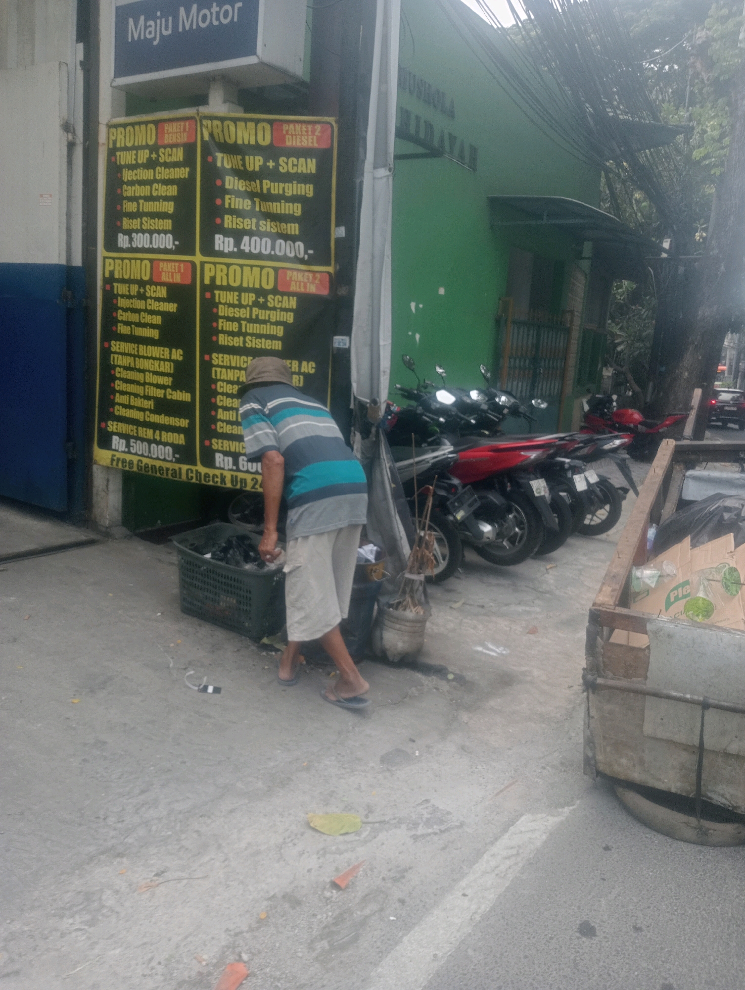 The image captures a street scene in South Jakarta, Indonesia, near a motorcycle repair shop. In the foreground, a man wearing a striped t-shirt and light-colored shorts is sorting through a black plastic basket filled with various items, possibly for recycling or disposal. He is wearing flip-flops and a tan hat. To his left, a motorcycle repair shop displays promotional posters with services like "TUNE UP + SCAN," "Injection Cleaner," "Carbon Clean," and "Fine Tuning," with prices ranging from Rp. 300.000 to Rp. 500.000. Behind the man, several motorcycles are parked. The environment is a mix of urban infrastructure with a green building in the background, a sidewalk, and overhead electrical wires. There are also some potted plants and a trash bin.